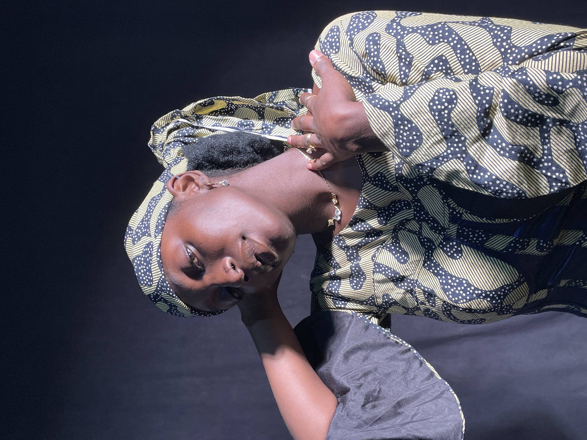 The image is a portrait of a person, likely an adult woman, with dark skin, positioned against a plain, dark background. She is wearing a headwrap and garment made from a distinctive patterned fabric, featuring black/dark blue areas with light yellow/gold stripes and white dots. Her dark, curly hair is visible beneath the headwrap.
The subject's head is tilted significantly to her right (viewer's left), and her gaze is directed towards the camera with a calm, slightly contemplative expression. Her mouth is slightly open. She wears small earrings, a delicate necklace, and rings on her fingers. One hand is gently placed near her head/neck area, and her other arm, with a sleeve made of a solid dark fabric, is visible lower down.
The lighting appears to be artificial, highlighting her features and the vibrant patterns of her attire, while casting soft shadows. The overall composition is a posed shot, focusing on the individual and her unique style. There is no visible text in the image. The location context provided is Jos, Nigeria.