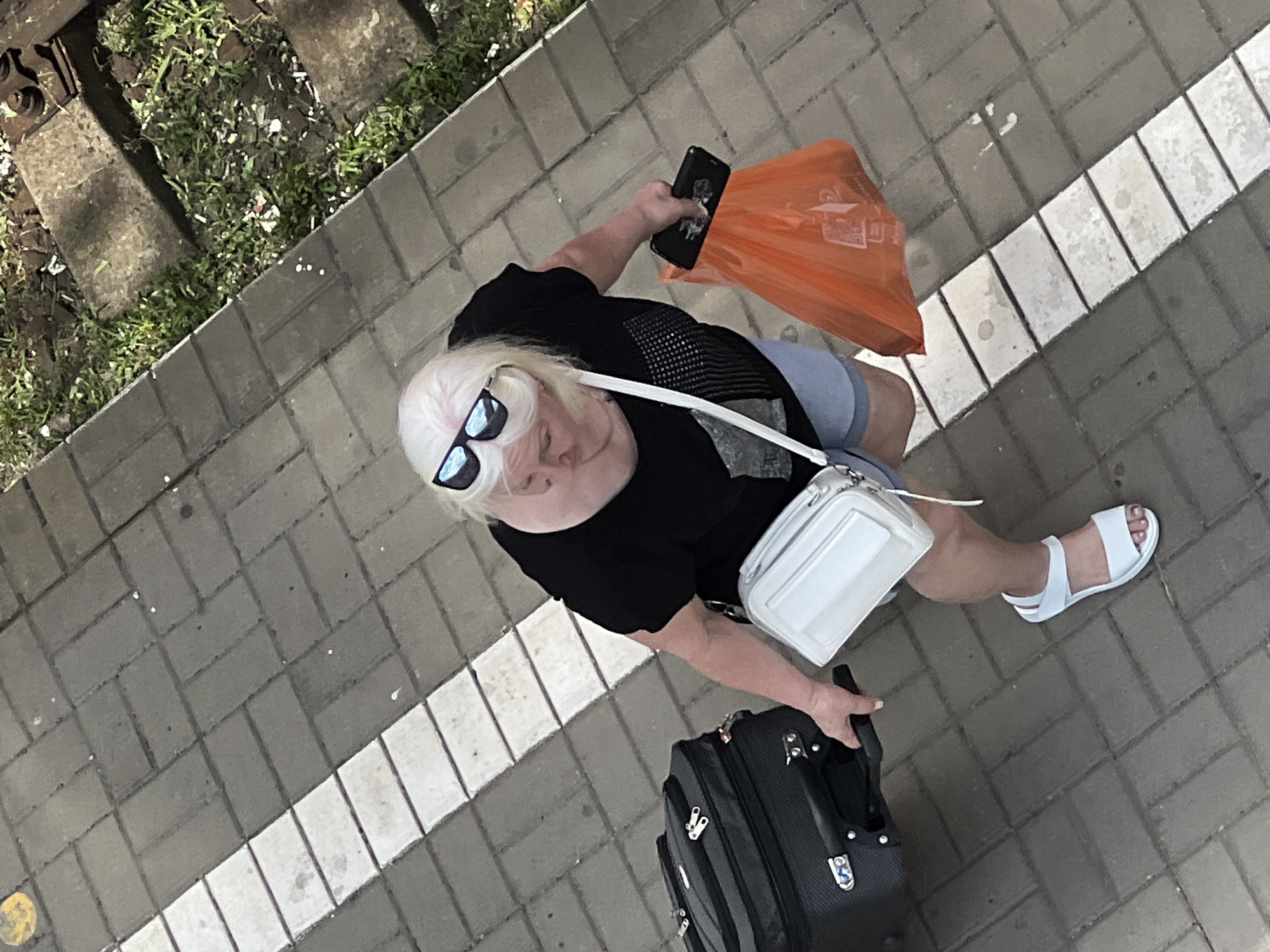 A person with blonde hair and sunglasses is walking on a paved walkway. They are wearing a black t-shirt and light-colored shorts. A white purse is slung across their body. They are holding a black smartphone in one hand and an orange shopping bag in the other. A black suitcase with wheels is being pulled behind them. The walkway is made of gray paving stones and has a white line painted on it. To the left of the walkway, there is an area with grass and what appear to be railway tracks or a similar industrial setting. The lighting suggests it is daytime, possibly sunny given the sunglasses. The person's posture and the luggage indicate they might be traveling or on their way somewhere. There is no visible text in the image. The setting appears to be outdoors in an urban or semi-urban environment, possibly near a train station or a similar transit point, given the proximity to what looks like train tracks.