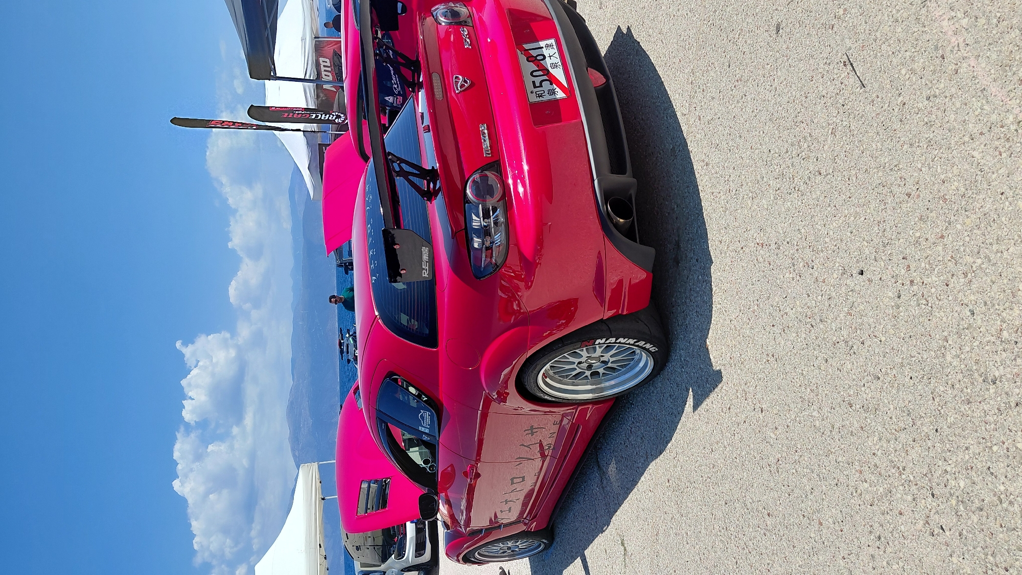 In the Municipal Unit of Aegio, Greece, a bright pink Mazda RX-8 is parked on a paved surface, likely at an outdoor event. The car features a prominent rear spoiler, custom wheels with white-lettered tires, and Japanese characters on its side and rear windshield. A license plate with the characters "50-81" is visible on the back. In the background, a body of water, possibly the sea, is visible, with a mountainous landscape and a clear blue sky with scattered clouds. Several white tents and tall black banners are also present, suggesting a car show or exhibition. A man in a dark green shirt stands near the water.