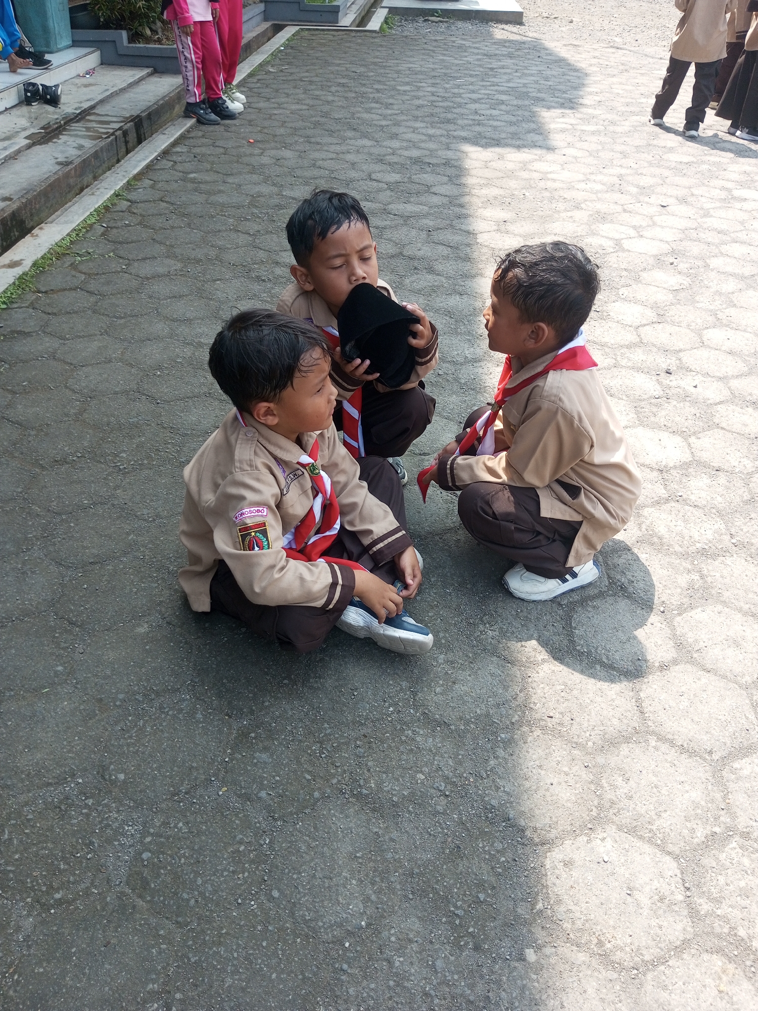 Three young boys in scout uniforms are sitting on a paved outdoor surface. The boy on the left is looking to his right, holding a black object in his hands. The boy in the middle is looking downwards, also holding the black object. The boy on the right is looking to his left, with his hands in his lap. The ground is paved with hexagonal concrete blocks and has scattered gravel. Sunlight creates distinct shadows on the ground. In the background, there are stairs with plants and a glimpse of a blue structure. The scene appears to be in an outdoor area, possibly a schoolyard or community space, consistent with the description of the city of Kalibeber, Indonesia.