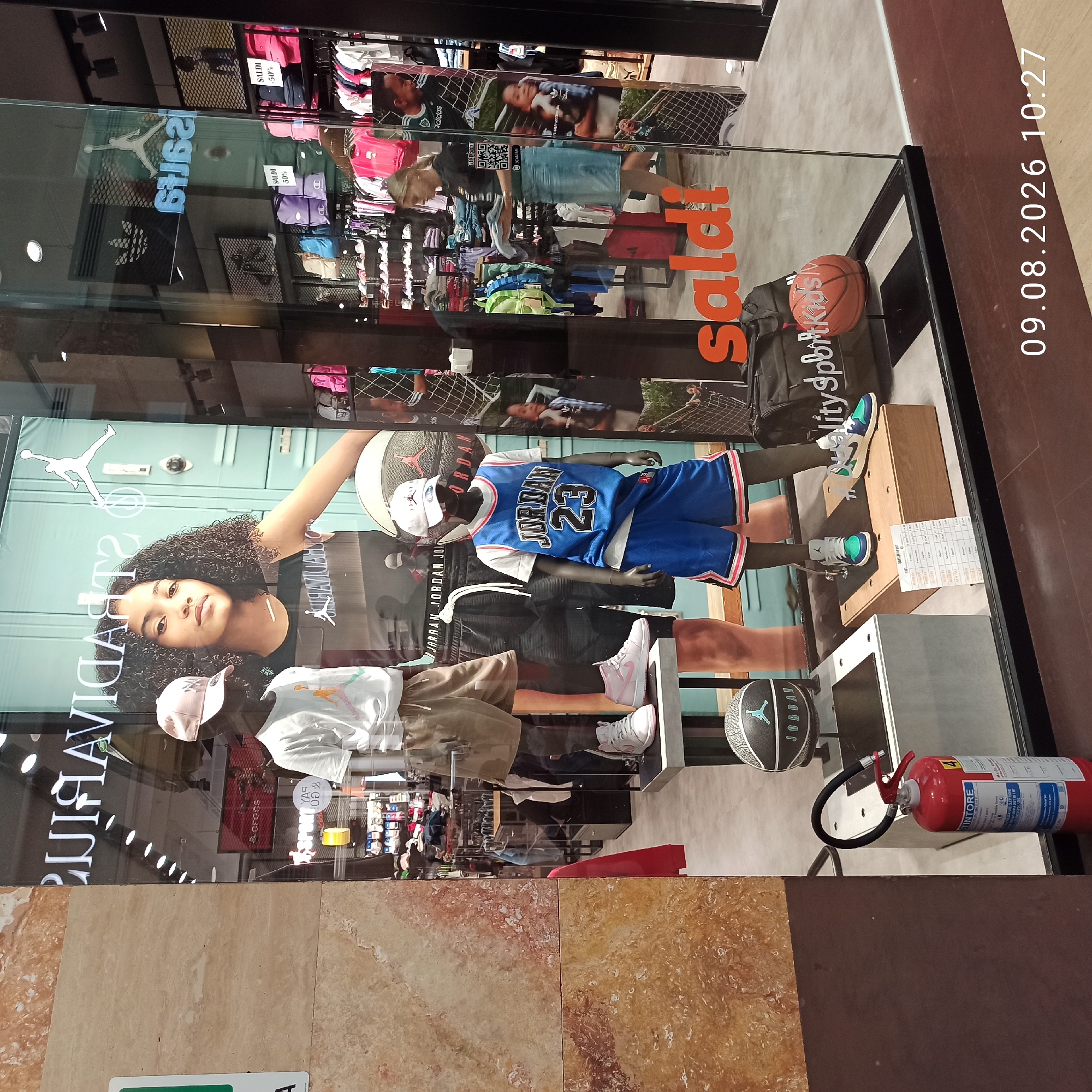 This is a street-level, eye-level, medium shot of a storefront display window in Molfetta, Italy, dated 09.08.2026 at 10:27 AM. The display showcases athletic apparel and footwear, primarily featuring the Jordan brand. Two mannequins are dressed in Jordan-branded clothing: one in a white t-shirt and khaki shorts, and another in a blue Jordan basketball jersey with the number 23 and matching blue shorts. A pink baseball cap is placed on the head of the first mannequin, and a white Jordan cap sits on the second. A basketball with the Jordan logo is positioned on a stand. Adjacent to the mannequins, there is a red fire extinguisher. The display also includes a large promotional poster of a young Black girl with curly hair wearing a Jordan tank top. In the background, within the store, shelves are stocked with various colorful backpacks, sneakers, and clothing items. A woman is visible inside the store, handling some clothing. Large signage on the window reads "saldi" in orange lettering, indicating a sale. Reflections in the glass show other store displays and architectural elements. The overall environment suggests a retail setting focused on sports fashion.