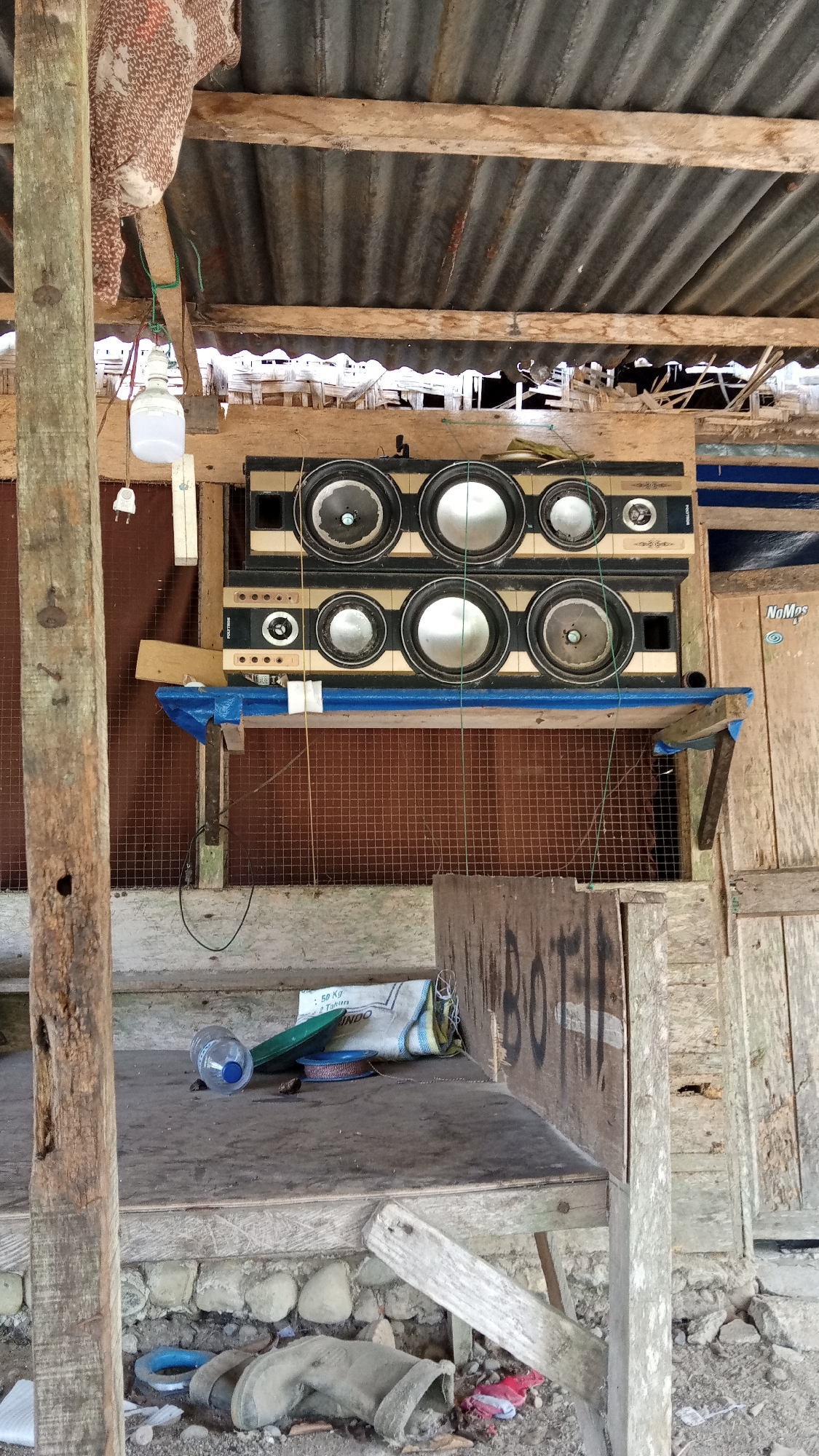 The image depicts an outdoor stall or structure in Kebonbaru, Indonesia. The stall has a corrugated metal roof supported by wooden beams. Below the roof, a wooden shelf holds a large speaker system composed of multiple circular speakers arranged in two rows. A blue tarp is draped over a wooden support beneath the speakers. In front of the speaker system, on a wooden surface, lies a plastic bottle, a spool of wire, and a rolled-up bag. The stall is constructed with rough wooden planks and a wire mesh backing. In the foreground, the ground is littered with debris, including rocks, discarded boots, and plastic items. Graffiti reading "BOTI" is visible on a wooden panel to the right. The overall scene appears to be an improvised outdoor setup, possibly for selling goods or providing entertainment.