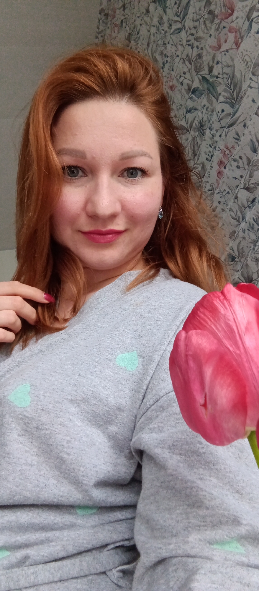 A medium shot depicts a fair-skinned woman with red hair, blue eyes, and pink lipstick, wearing a gray long-sleeved shirt with light green heart patterns. She is smiling slightly and looking at the camera. Her right hand is near her neck, with a pink-painted fingernail visible on her index finger. To the right of the frame, a partially visible pink tulip is positioned. The background shows a wall with a floral pattern in shades of green and pink. The location appears to be indoors in a city and state of Unknown.