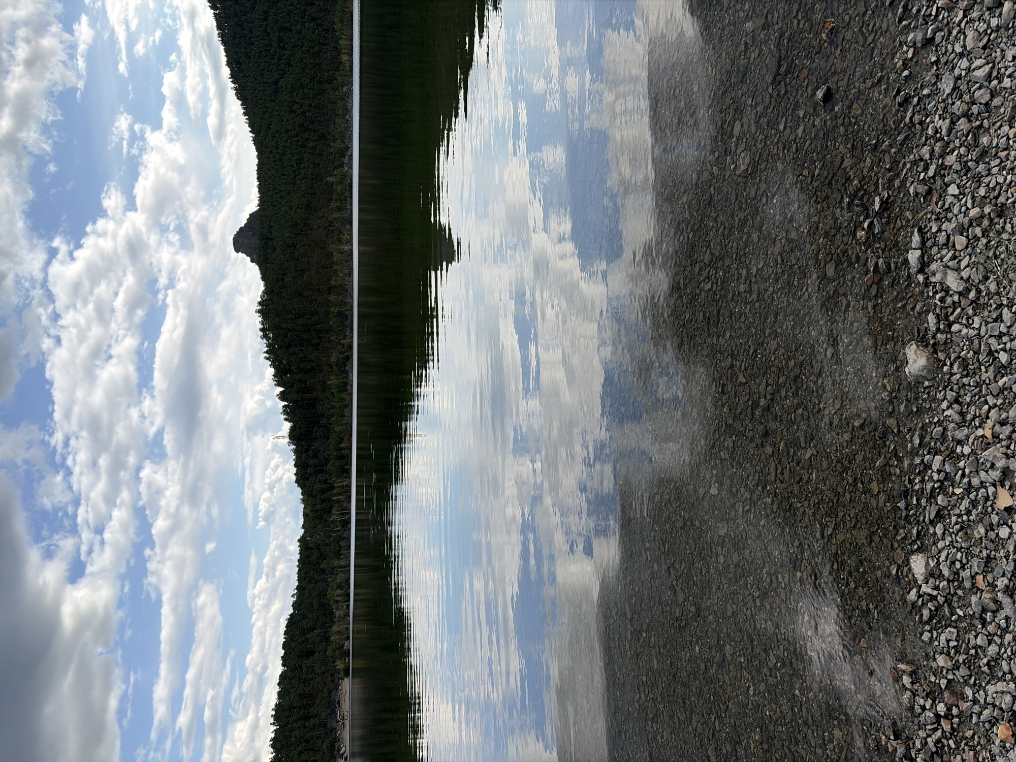 The image depicts a serene natural landscape. In the foreground, a rocky shoreline transitions into a calm body of water. The water's surface is smooth, reflecting the sky and surrounding trees with remarkable clarity, creating a mirror-like effect. Beyond the water, a dense forest of dark green trees covers the hills that form the horizon. A prominent rocky peak, resembling a distinct formation, stands out against the treeline on the right side of the image. The sky above is a mix of blue and white clouds, suggesting an overcast but pleasant day. The overall scene evokes a sense of tranquility and natural beauty. The location is identified as Зелёный Бор ауылдық округі, Kazakhstan. There are no people or specific man-made structures visible in the image, emphasizing the untouched nature of the environment.