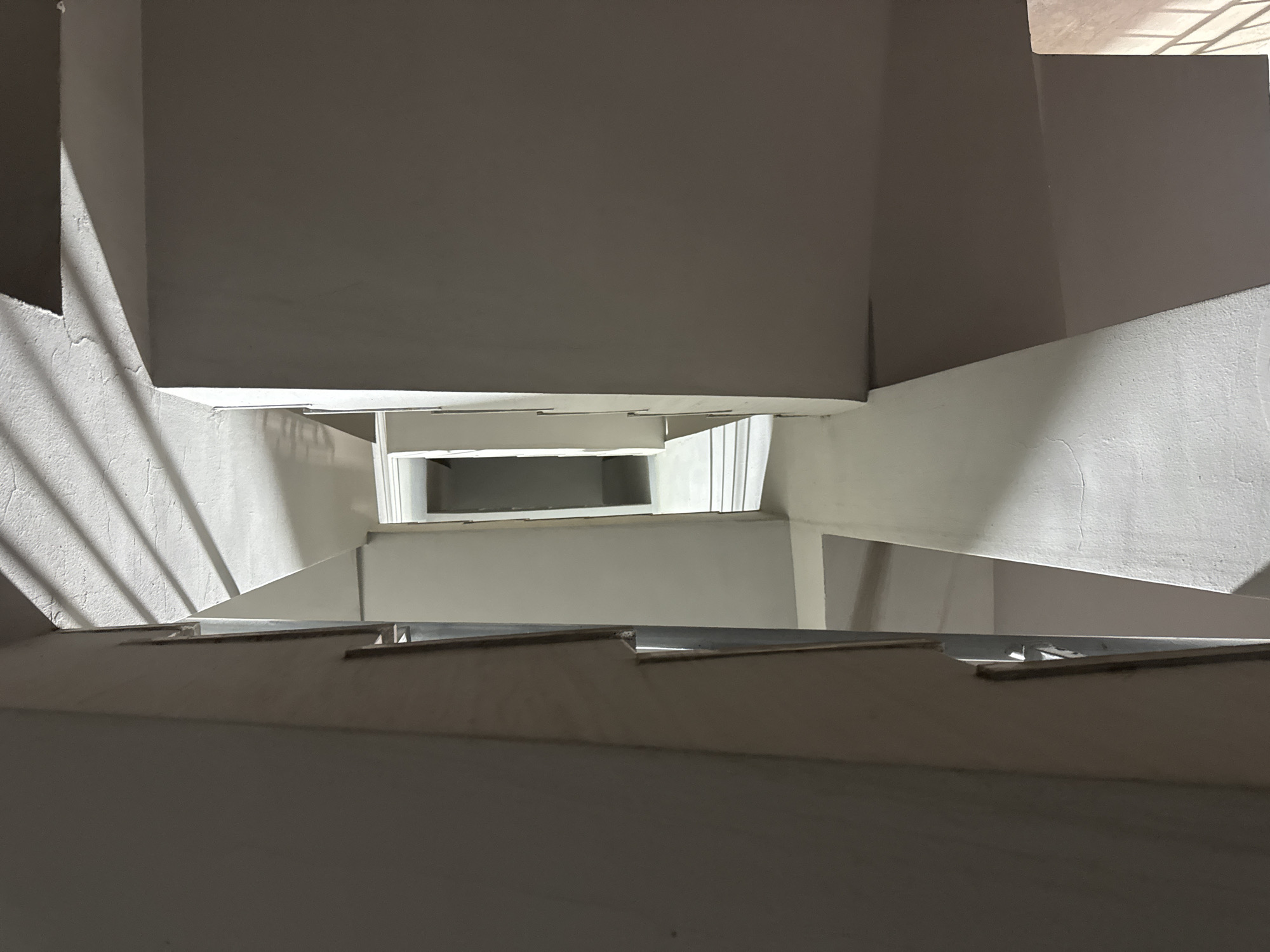 The image depicts a top-down view of a white stairwell. Light streams in from a window on the upper right. The stairwell is enclosed by gray walls. The steps are evenly spaced. No people or objects are visible in the stairwell. No location information is included in the original image. Therefore, I cannot determine if this image is in Yerevan, Armenia.