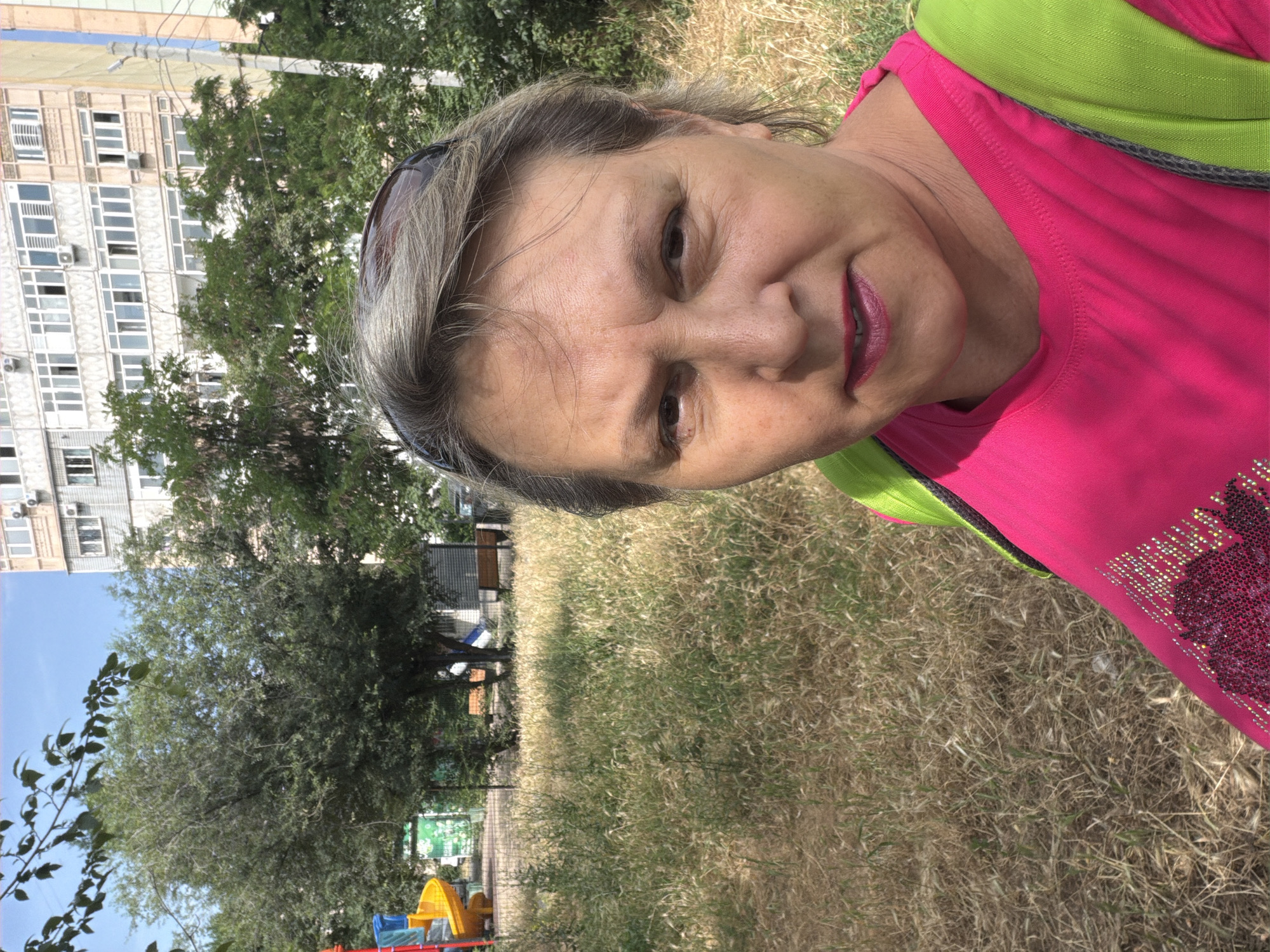 A middle-aged woman with graying hair and sunglasses perched on her head stands outdoors, facing the camera. She is wearing a bright pink t-shirt with decorative sequins and a lime green backpack. Her expression is neutral. The background consists of dry, yellowed grass, some trees, and a tall, multi-story building with many windows. To the left, a colorful playground structure is visible. The scene appears to be in a residential area, possibly a park or courtyard.