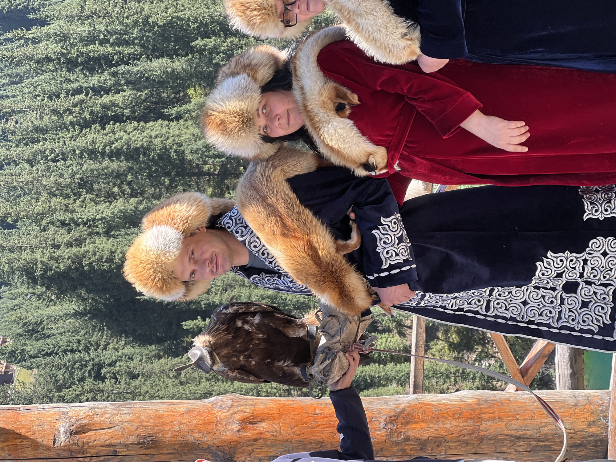 In the city of Kurmeti, Kazakhstan, on a sunny day, a group of people are gathered outdoors, with a backdrop of a dense, green forest. In the foreground, a man and two women, dressed in traditional fur-trimmed robes and hats, are posing for a photo. The man, with a stern expression, wears a dark robe with intricate white embroidery. He holds a large, brown eagle on his gloved hand, its talons gripping a leather perch. The woman to his right, in a vibrant red velvet robe and a similar fur hat, looks towards the camera. Another woman, partially visible on the far right, also wears traditional attire. To the left, a person's arm is visible, also wearing a dark sleeve, and their hand is extended, seemingly assisting with the eagle. A thick, wooden post stands to the left, adding to the rustic setting. The overall scene suggests a cultural demonstration or tourist experience involving falconry.