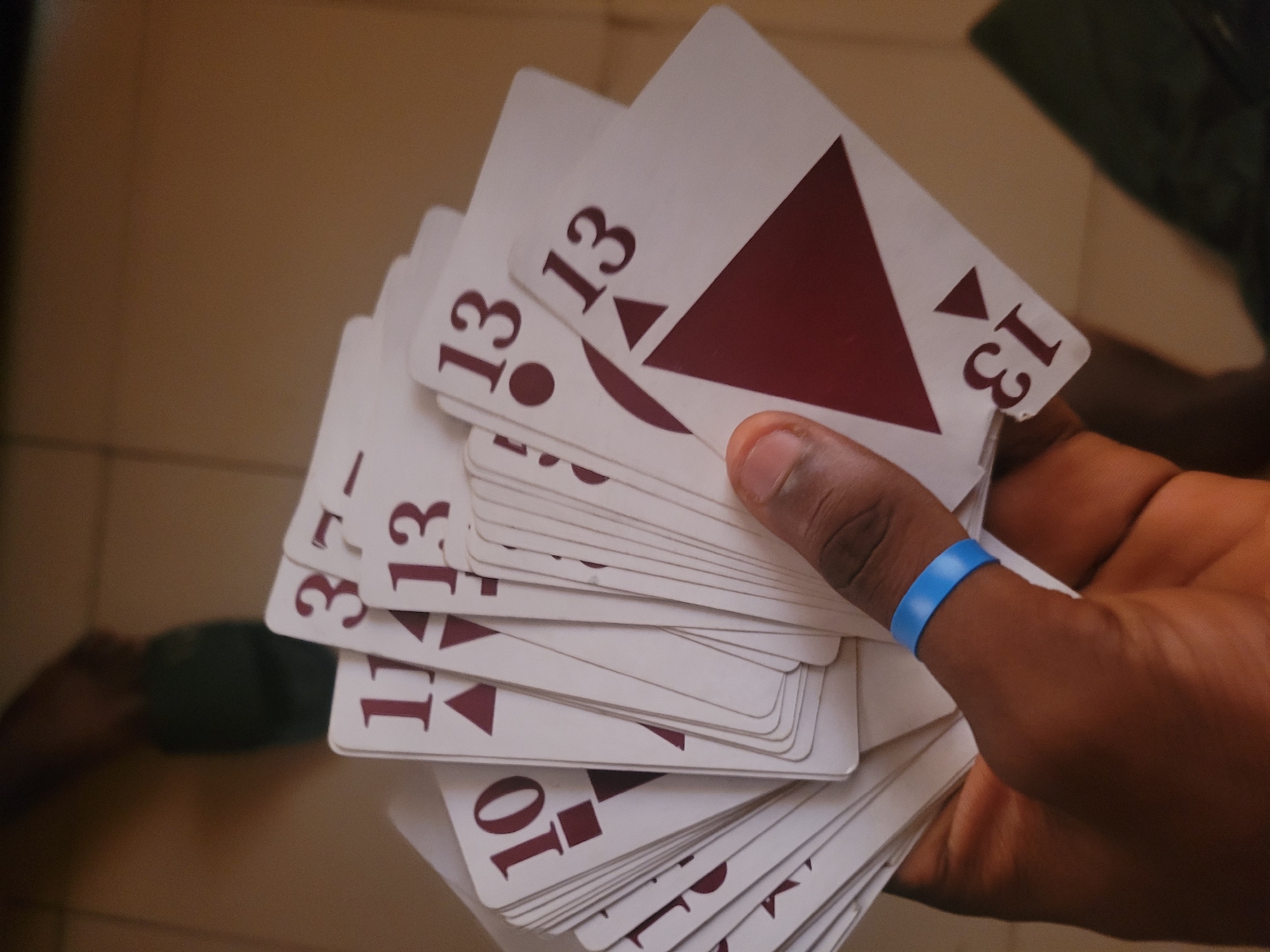 A close-up, high-angle shot shows a person's hand holding a fanned-out deck of cards, with their forearm visible. The hand is adorned with a bright blue wristband, and the cards, predominantly white with maroon markings, display various numbers and geometric shapes. The visible numbers include "10", "11", "13", and "37". The background is blurred, suggesting an indoor environment with tiled flooring and hints of a person's legs in dark green trousers. The lighting is soft, casting subtle shadows.