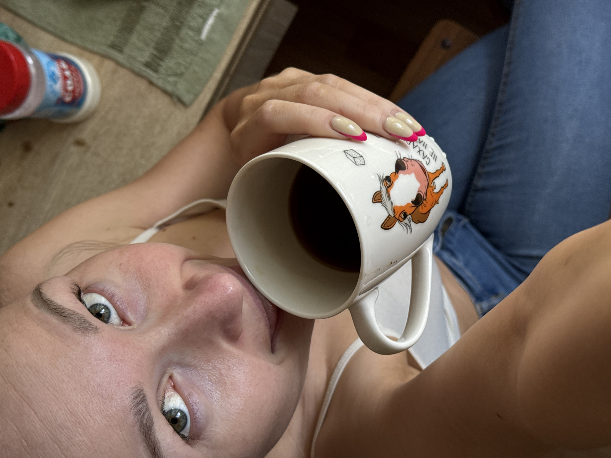 A woman in a white top and blue jeans is taking a selfie while holding a white mug with a cartoon character on it. The mug contains a dark liquid, possibly coffee. She is indoors, as indicated by the interior setting and the presence of household items like a bottle of cleaning supplies and a towel in the background. The location is not identifiable from the image.