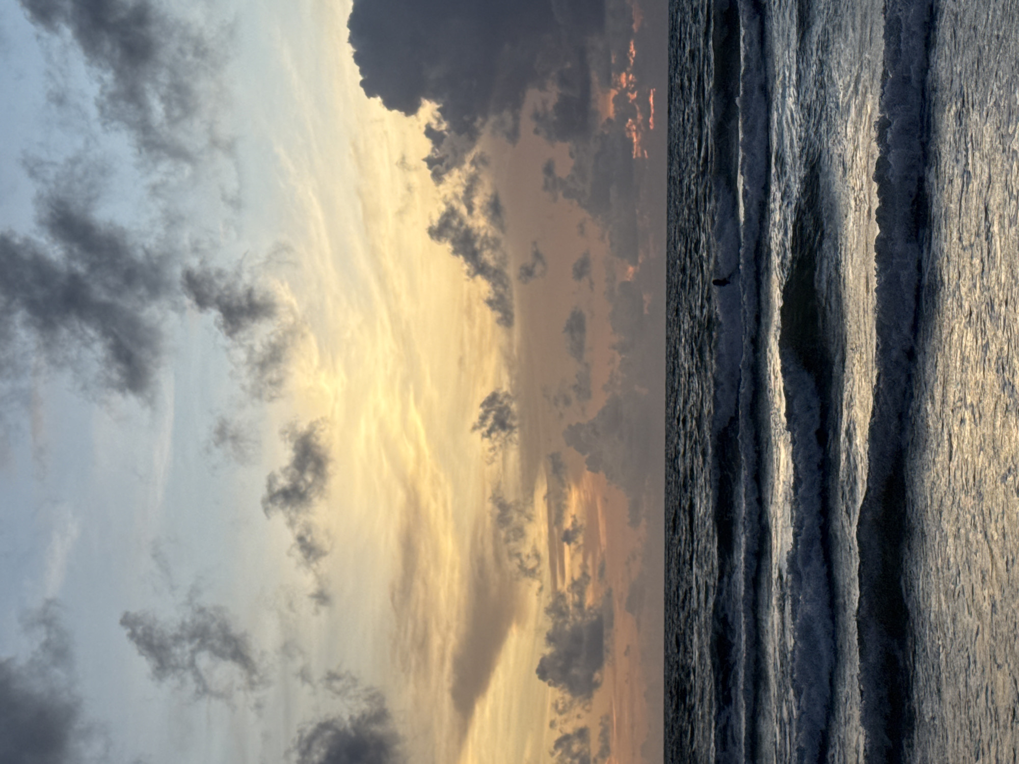 The image depicts a seascape at sunset over the ocean in Patong, Thailand. The sky is a gradient of pale blue and soft yellow, with scattered grey clouds. The sea is dark, with visible waves breaking and creating white foam. In the middle ground, a lone surfer is visible as a dark silhouette against the breaking waves, appearing to be paddling or riding a wave. The lighting suggests the sun is low on the horizon, casting a warm glow on the clouds and reflecting on the water's surface. The overall atmosphere is calm and serene.