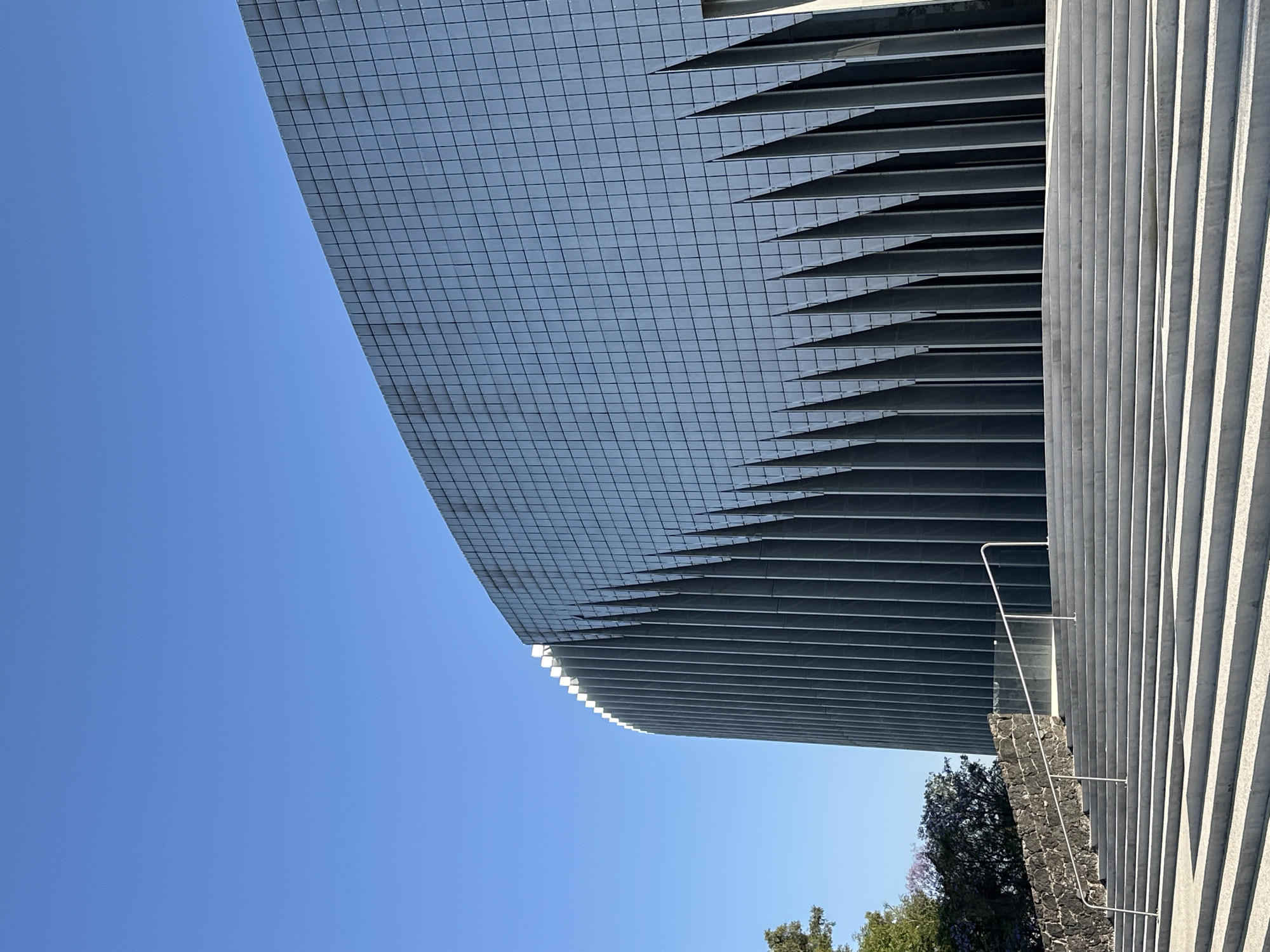The image shows a portion of the Soumaya Museum in Mexico City, Mexico, viewed from a low angle. The museum's distinctive, curved façade of hexagonal aluminum tiles is visible against a clear blue sky. To the right, there are vertical rows of stepped, bright stone, likely the museum's exterior or a nearby structure. The corner of the museum is lined with horizontal stone beams to give it a tiered and segmented architectural look.