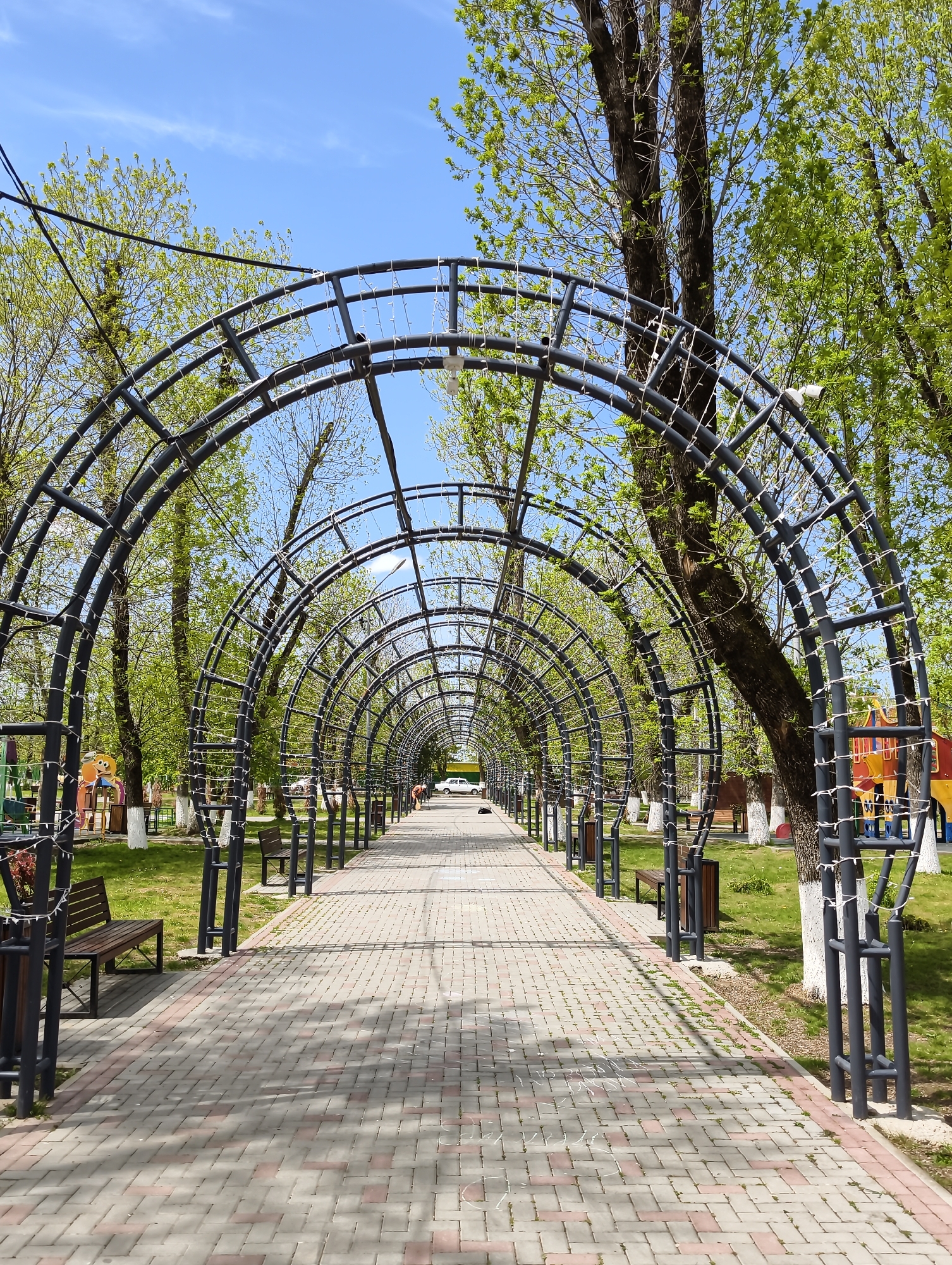 The image displays a picturesque park pathway in Ilskiy, Russia, captured on a bright, sunny day, likely during spring due to the fresh, light green foliage on the trees. The central feature is a long, tunnel-like structure made of dark grey/black metal archways that span the width of the path. These arches are decorated with strings of small white lights or cabling, possibly for evening illumination or as supports for climbing plants.
The pathway itself is paved with light grey and reddish rectangular pavers arranged in a herringbone pattern, leading the eye deep into the scene. On either side of the path, lush green grass stretches towards rows of deciduous trees, many with their lower trunks painted white. Wooden park benches with dark frames are positioned at regular intervals on the grassy areas, inviting visitors to rest.
In the background, colorful playground equipment, including slides and climbing structures, is visible on both sides of the path, suggesting a family-friendly recreational area. A white security camera is mounted on one of the archways on the right. In the far distance, a light-colored vehicle and a faint human figure can be seen at the end of the path. The sky above is a clear, vibrant blue with a few scattered, wispy white clouds. No discernible text is visible in the image.