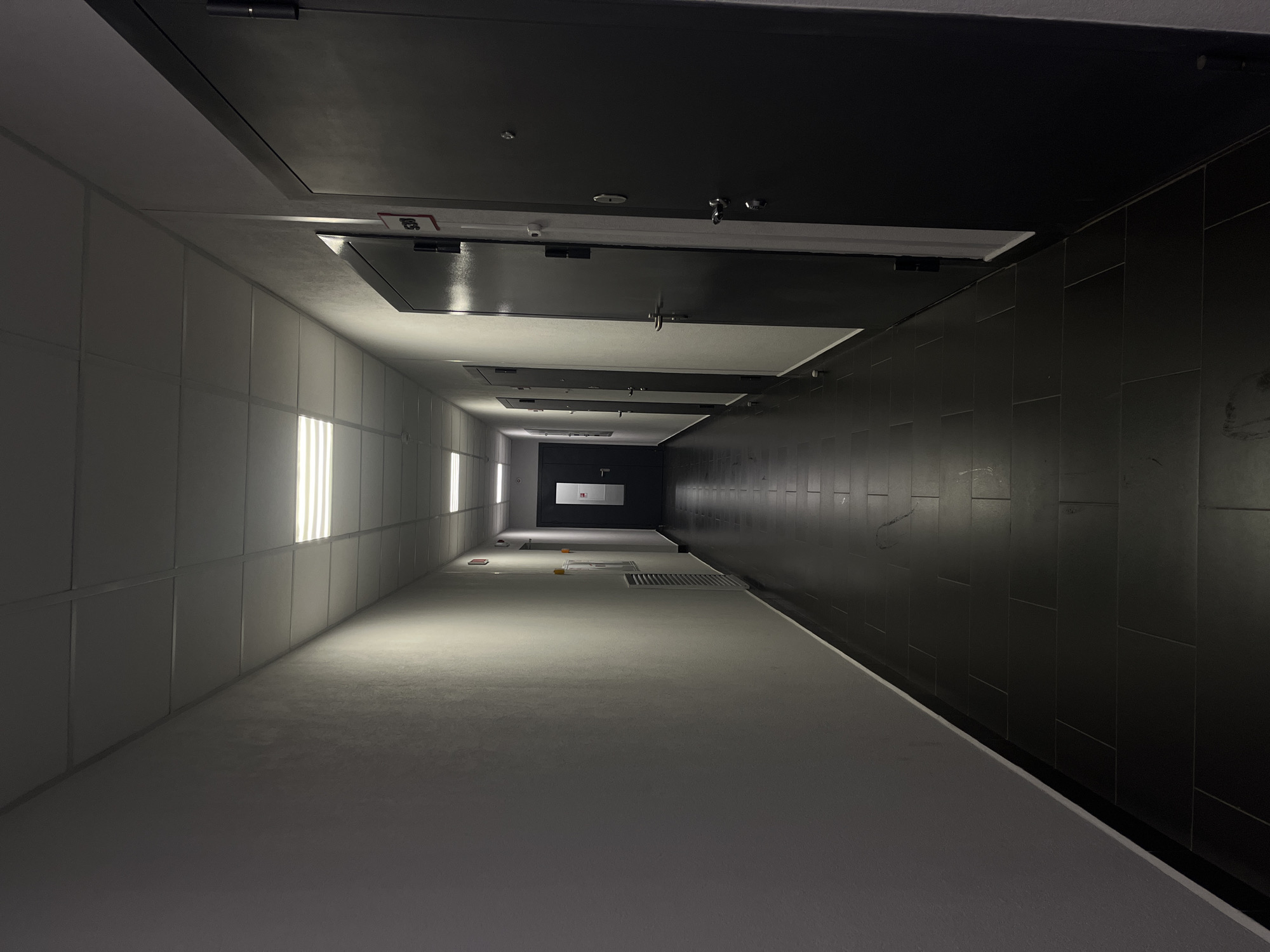 The image depicts a long, narrow hallway or corridor with dark tiling on one side and a white, textured surface on the other. Fluorescent lights illuminate the white side of the hallway. Dark doors are positioned above, creating a sense of enclosure. The perspective suggests the photograph was taken at a low angle, looking down the length of the corridor. The scene appears sterile and institutional. The location is not identifiable as Saint Petersburg, Russia based on visual features in this image.