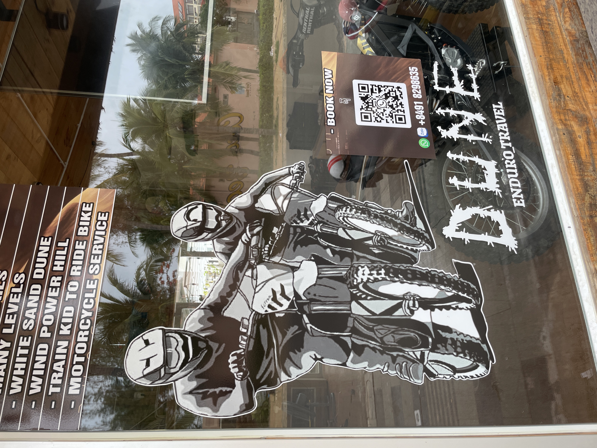 The image shows a window display in Phu Thuy Ward, Vietnam advertising "Dune Enduro Travel". The display includes information about motorcycle service, training, and tours, including "White Sand Dune" and "Wind Power Hill" options. There is a graphic of two people riding dirt bikes. A "Book Now" sign with a QR code and phone number is also visible. A dirt bike is visible through the window.