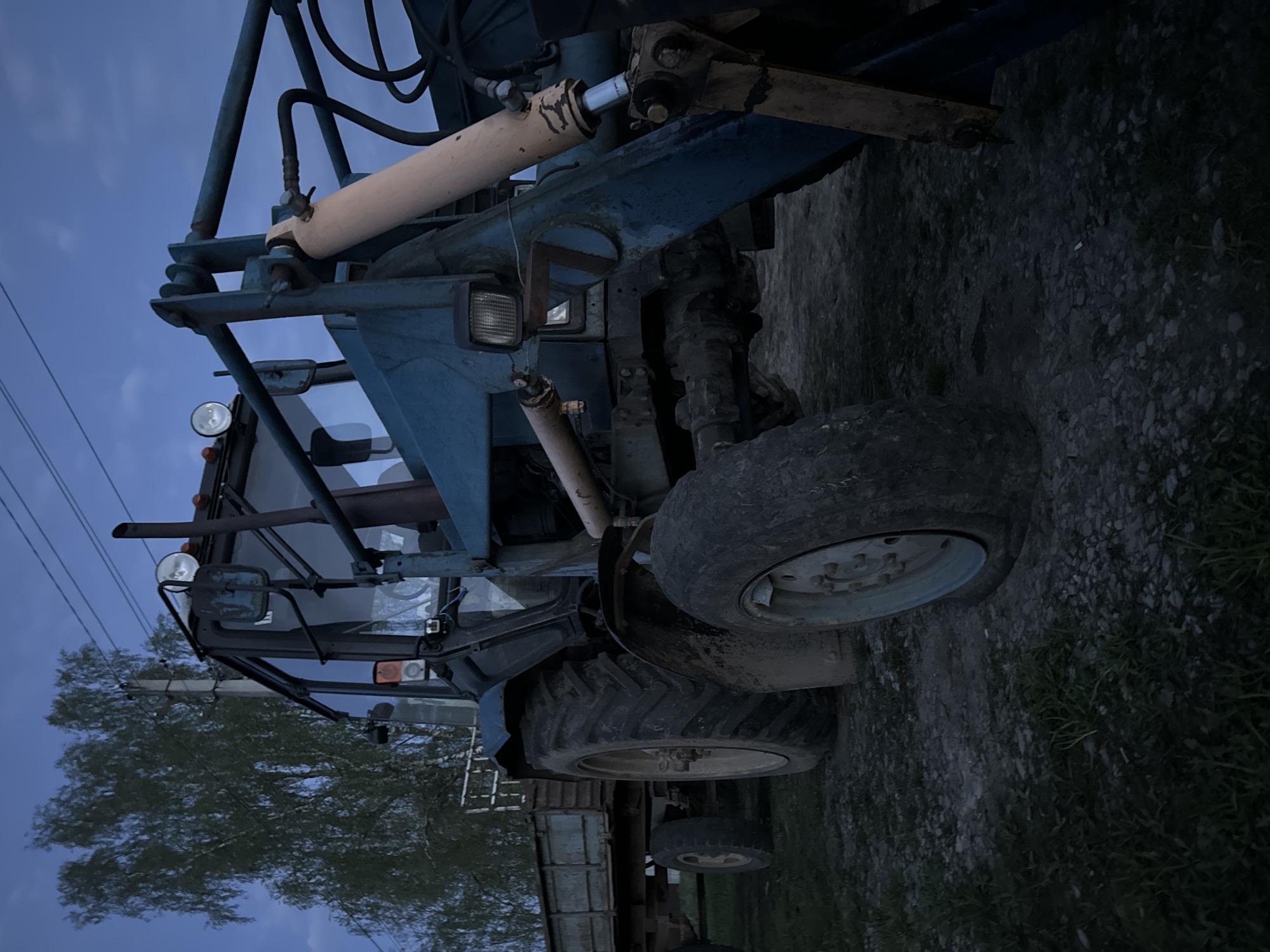 The image shows a blue tractor parked outdoors, possibly on a gravel or dirt surface. The scene appears to be near a wooded area or trees. The sky is visible, suggesting it might be dusk or dawn. There are power lines visible in the background. The tractor is likely located in Красный Маяк, Russia, as specified.