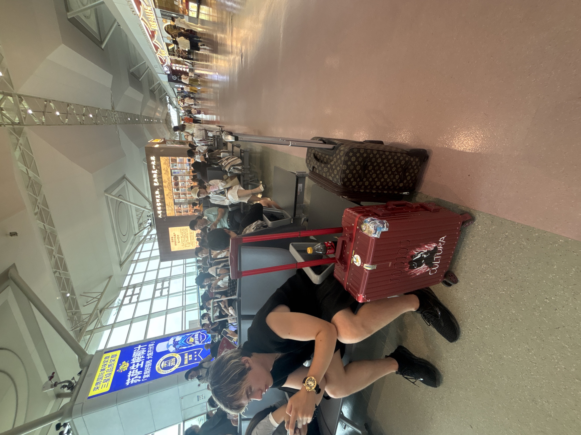 This is a medium shot, eye-level, full-frame photograph taken in an airport terminal. The foreground is dominated by a young man with blonde hair, wearing a black sleeveless shirt, black shorts, and black athletic shoes. He is seated on a gray airport bench, looking down at his mobile phone, and holding a second phone in his left hand. A gold watch is visible on his left wrist. To his right are two pieces of luggage: a red, rectangular suitcase with "CULTURA" written on it, and a brown, patterned suitcase. A bottle of Coca-Cola is propped against the red suitcase.
The background shows a busy airport waiting area. Numerous people are seated on similar gray benches, many also looking at their phones or engaging in conversation. The terminal features a high, modern ceiling with exposed metal beams and lighting fixtures. Large windows allow natural light to enter, illuminating the shiny, light-pink tiled floor. To the left of the frame, a large blue advertisement panel with Chinese text and graphics is mounted on a white pillar. Further down the terminal, shops and other advertisements are visible along the right side of the hallway. The overall environment suggests a public space, likely a departure lounge. The presence of the advertisement panel with Chinese text, and the general architectural style, indicate the location is likely within China. Based on the specific details of the advertisement, which mentions "Xinwu District" and "Wuxi" in Chinese text, the location is identified as the Xinwu District of Wuxi, China.