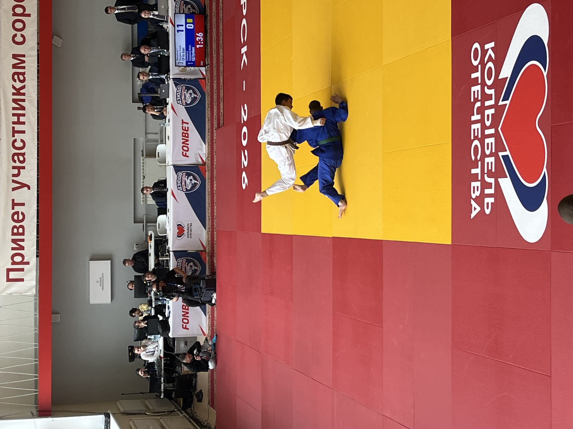 Here's a detailed description of the image:
**Scene:** An indoor judo competition. The scene is viewed from the side with the judging table in the distance.
**Subjects:** Two judokas are engaged in a match on a yellow and red judo mat. One judoka is wearing a white uniform with a brown belt, the other is in a blue uniform with a green belt.
**Objects:**
* Judo Mat: Two distinct colors, yellow and red, dividing the area.
* Banners: A banner with red text in Russian (partially visible, "Привет участникам сор") and another with "PCK - 2026". Logos are displayed on tables including FONBET.
* Judging Table: Several people are seated behind tables, presumably judges and officials. Digital scoreboards are visible on the table.
**Actions:** The two judokas are actively grappling, with the judoka in white appearing to have the upper hand.
**Interactions:** A judo match is in progress between two competitors. Officials at the judging table are observing.
**Environment:** The scene is indoors, presumably a sports arena or gymnasium. A wall is visible in the background. "FLY TECHNOLOGY" is inscribed on a box.
**Location:** The competition is sponsored in part by "Колыбель Отечества" or Cradle of the Fatherland and is held in Russia as denoted by the colors of the logo. Given the date, this location could be Krasnoyarsk in Russia, as it was the host of the 2026 Judo European Championships.