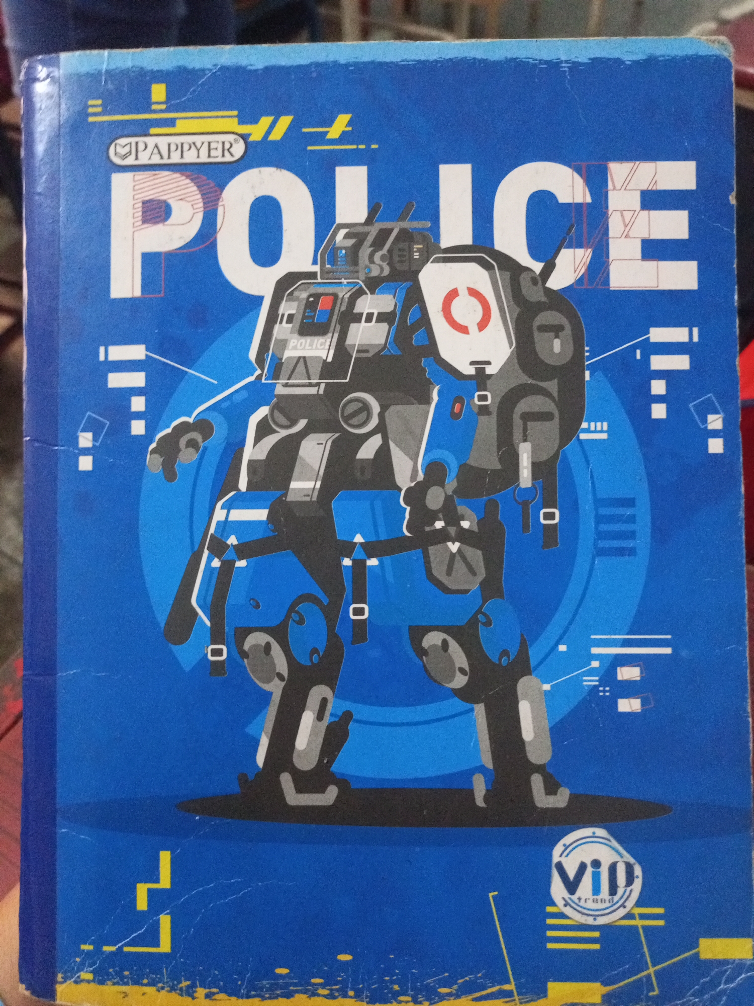 A blue notebook with a graphic of a robot wearing a police uniform is displayed. The robot is large and imposing, with a metallic gray and white design. The words "PAPPYER" and "POLICE" are printed on the notebook. The bottom right corner of the notebook features a circular logo with the words "VIP trend" inside. The notebook is lying flat, with its cover facing the viewer. The background is blurred, but appears to be an indoor setting. There is no indication of the location being Santa Ana, Venezuela.