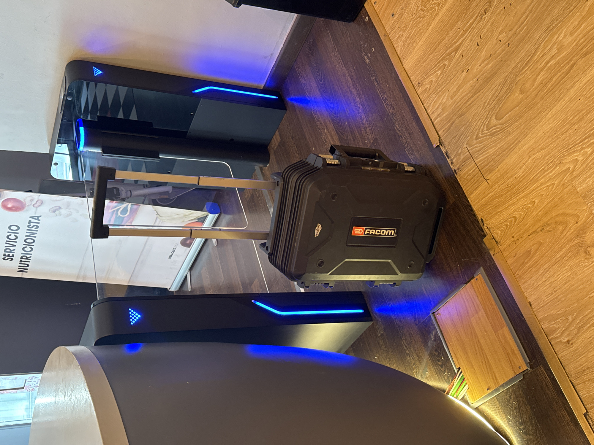 An overhead, slightly tilted shot depicts a black, rugged, wheeled case with a silver handle resting on a polished wooden floor. A blue light emanates from the right side of the case, casting a shadow. To the left of the case, a dark, modern-looking entrance gate with a blue light strip is visible. Further to the left, a curved, light-colored counter juts into the frame. Behind the gate, a large banner with Spanish text, likely advertising "SERVICIO NUTRICIONISTA," is displayed. The scene appears to be indoors, possibly in a commercial or public space, with the context of Murcia, Spain, suggested by the provided information.