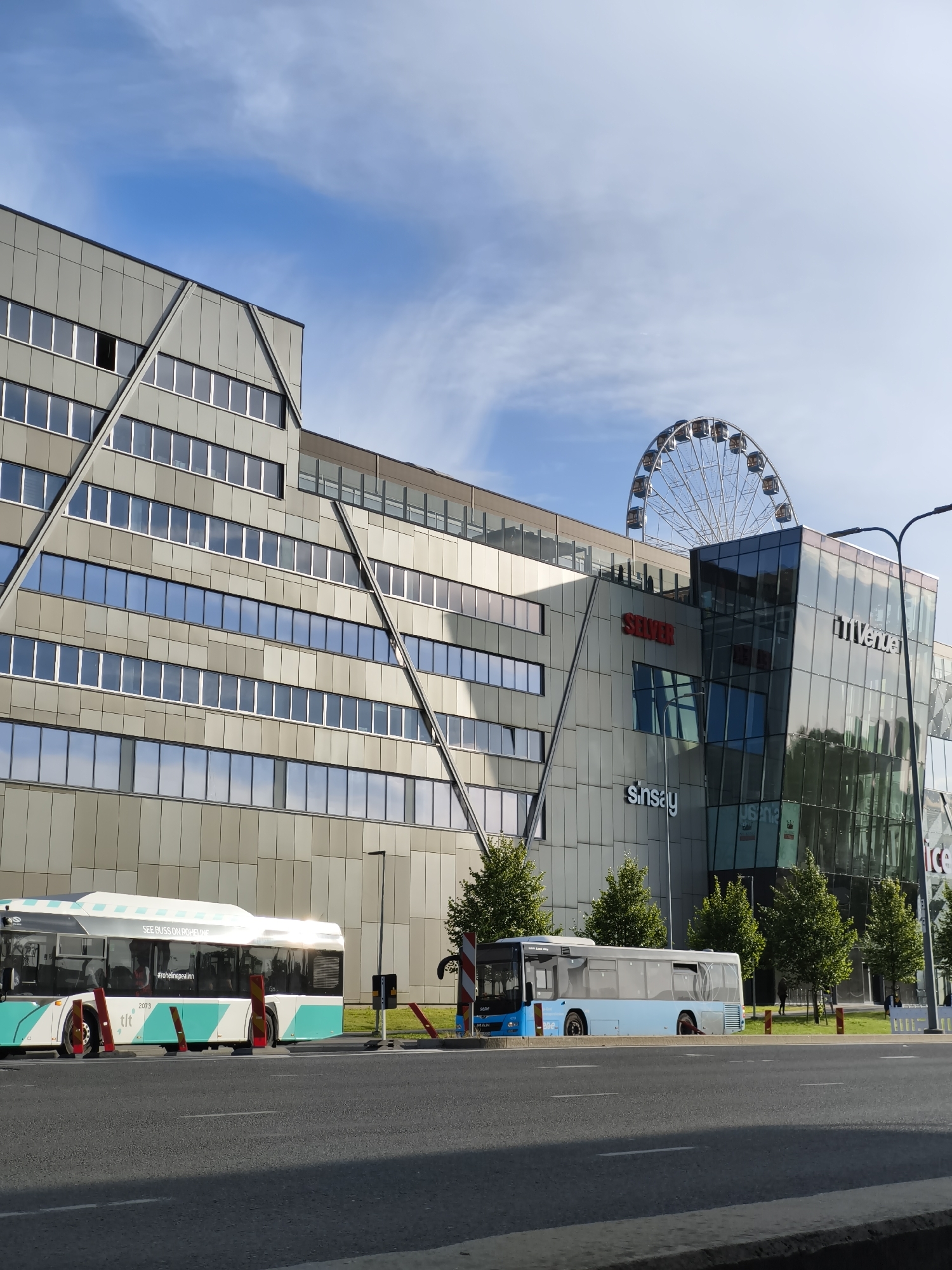 The image captures a street-level view of a modern building complex in Tallinn, Estonia, under a partly cloudy sky. The building features a striking angular design with a facade composed of grey metallic panels and numerous windows. A large Ferris wheel is visible behind the building.
In the foreground, two buses are parked along the roadside. The bus on the left is white with turquoise accents and displays text including "#rohelinepallinn". The bus on the right is light blue. The street in front of the buses is paved asphalt.
The building itself has a glass-fronted section with visible signage for "SEVER", "Sinsay", and "T1 Venue". The overall scene suggests an urban environment, likely a commercial or mixed-use area, with a focus on modern architecture and public transportation.