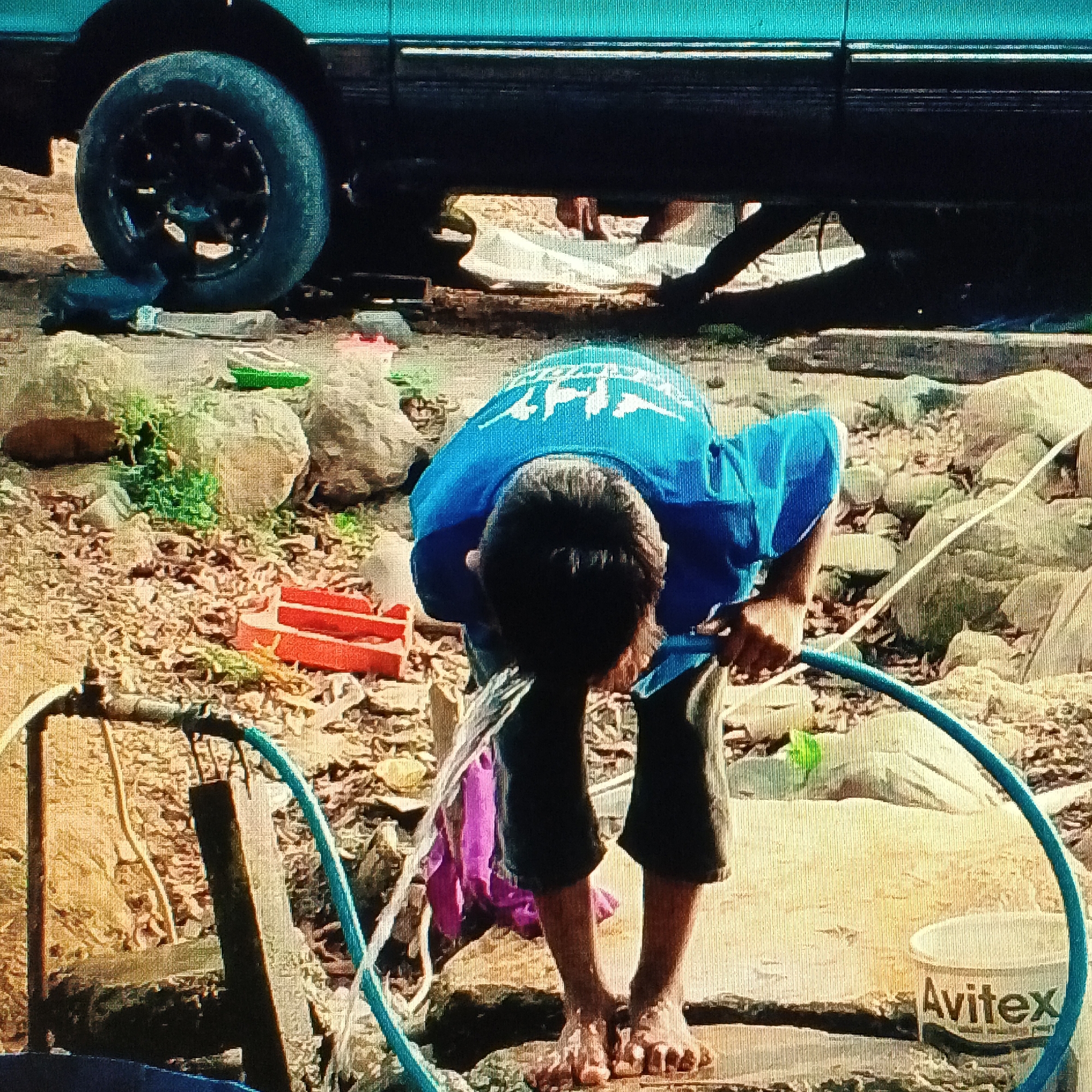 In Ende, Indonesia, a child wearing a blue t-shirt and dark shorts bends over a hose, appearing to be washing or playing with water. The child's head is obscured by their bent posture, but their hands are visible holding the hose. Water streams from the hose onto the ground. The scene is outdoors, with a tire of a vehicle visible in the upper left corner. The ground is covered with rocks, leaves, and some scattered plastic objects, including a red container and a green object. A white bucket with the label "Avitex" is present in the lower right. The overall environment suggests a rustic or informal outdoor setting.