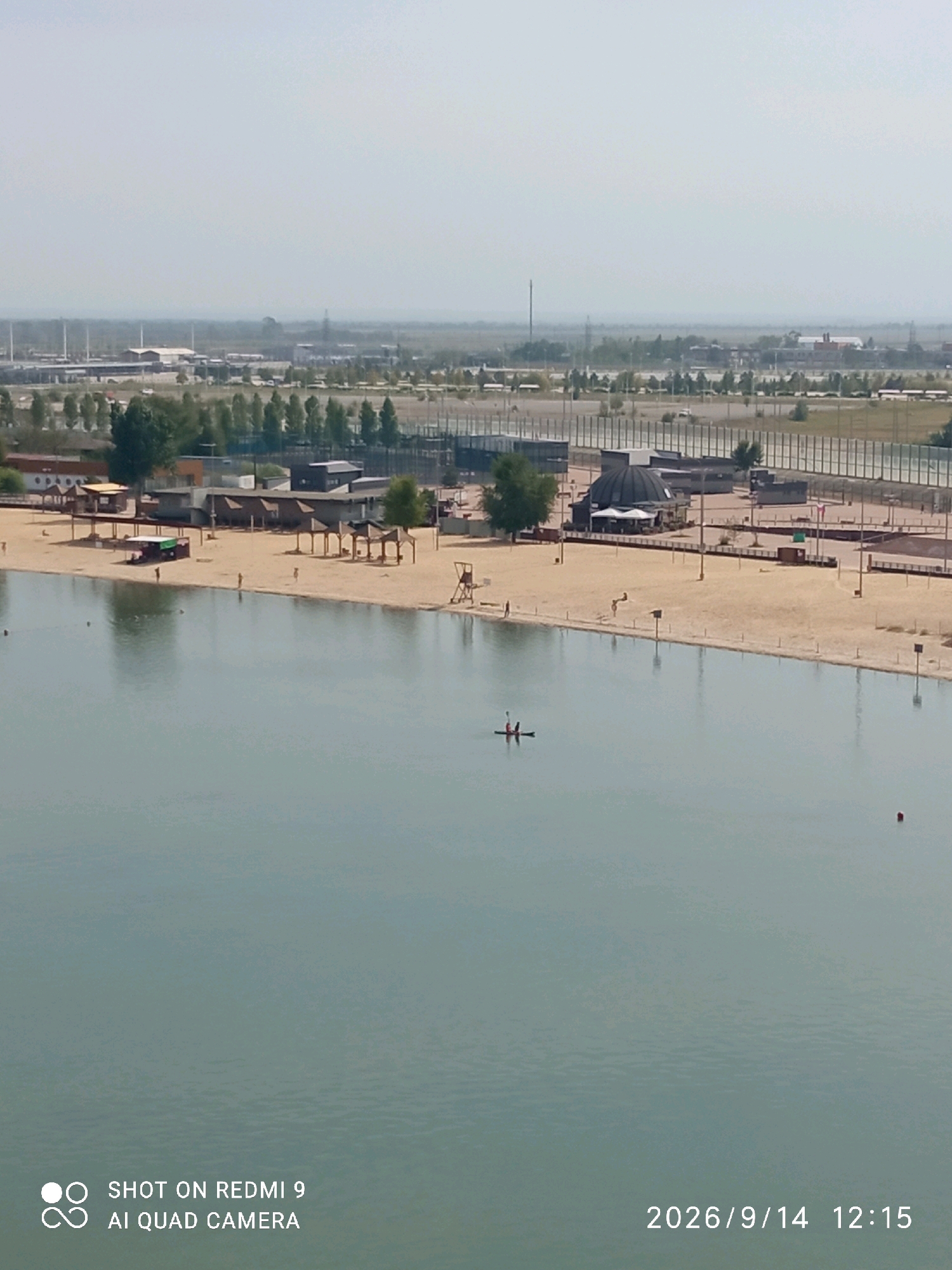 The image captures a wide shot of a man-made beach and lake area, likely within the city of Rostov-on-Don, Russia, given the context provided. In the foreground, a calm, light blue body of water occupies the majority of the frame. A person is visible in a kayak, paddling on the water. Scattered throughout the lake are a few small, dark buoys.
The middle ground features a sandy beach lined with a few small structures, likely cabanas or kiosks, offering shade. Several individuals are dispersed across the beach, some appearing to be standing and others walking. Further back, beyond the beach, is a flat, open area with some sparse vegetation and trees. In the distance, industrial buildings and structures are visible, suggesting the proximity of an industrial zone. The sky is overcast and hazy, creating a muted and diffused light across the scene. The image is timestamped with "2026/9/14 12:15" and indicates it was taken with a "SHOT ON REDMI 9 AI QUAD CAMERA".