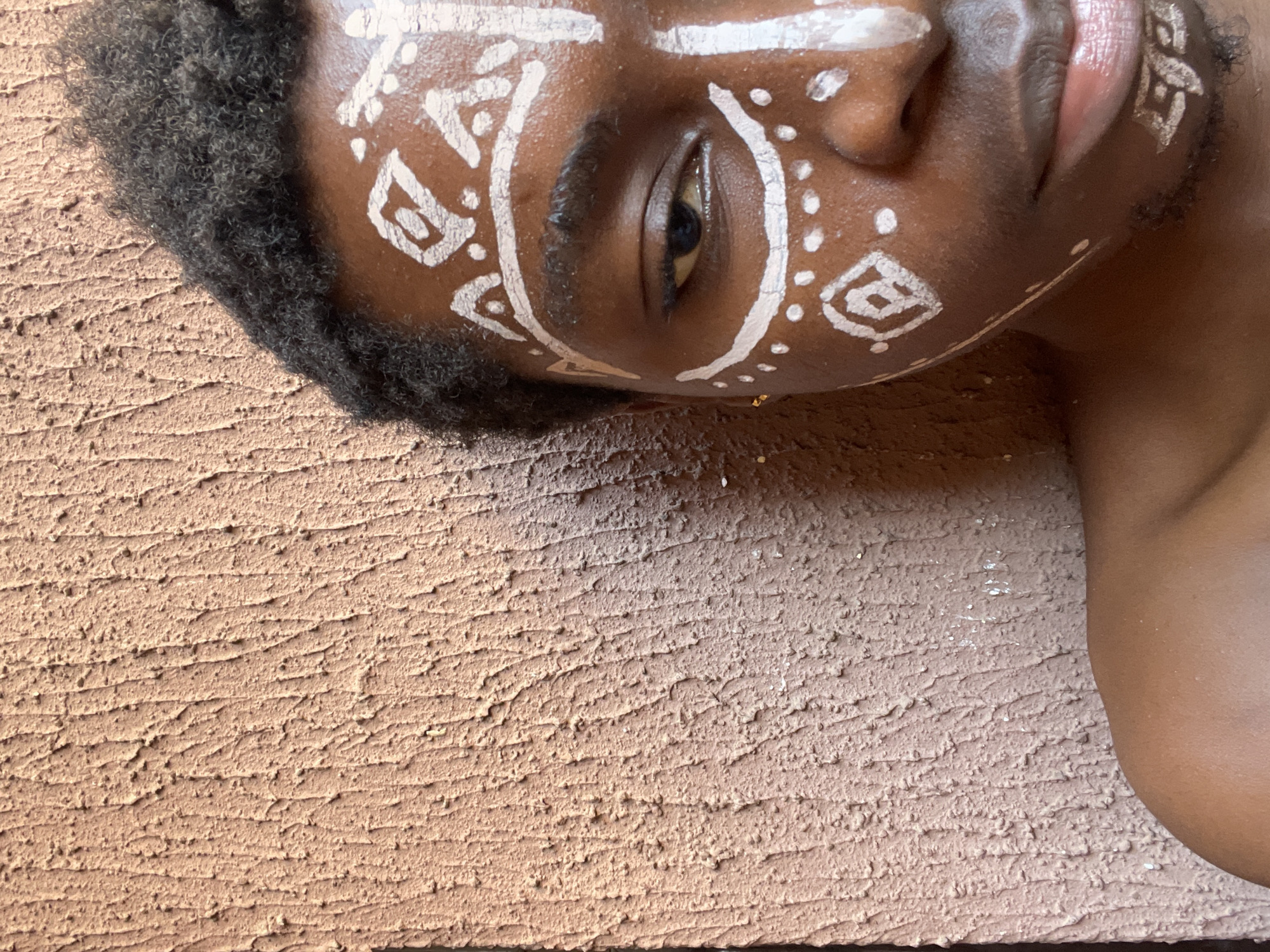 A close-up, high-angle shot depicts a dark-skinned person with geometric white paint designs on their face, lying on a textured, light brown surface. The person has dark curly hair and is partially visible from the chest up. The person's right eye is open, and their lips are slightly parted. The textured surface fills most of the frame. The location, Nkpor, Nigeria, is provided without specific contextual details within the image itself.