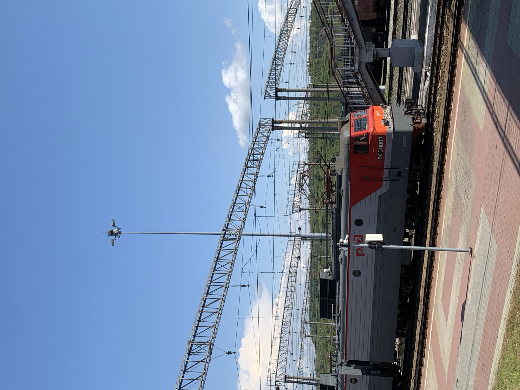 A train station platform in Prokopyevsk, Russia, with an electric locomotive. The locomotive is red and gray with Russian text and a number 8101007 on its side. It is on train tracks with overhead power lines. A footbridge is visible on the right, leading over the tracks. The platform is paved with red and gray tiles, and there is some green grass on the left. The sky is blue with some white clouds.
