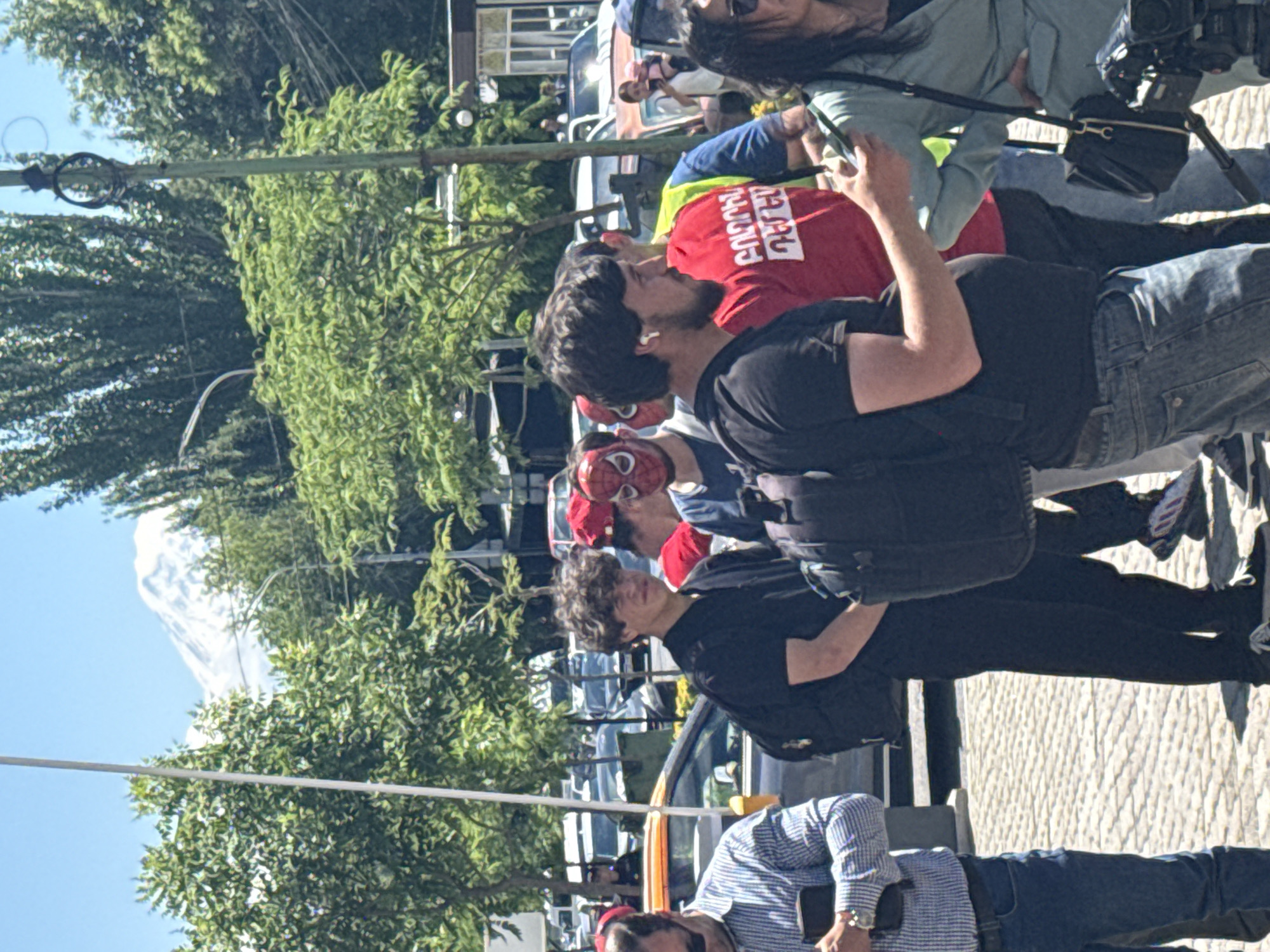 The image captures a group of people gathered outdoors on a sunny day. A tall mountain with snow can be seen in the distance. The location is likely in Yerevan, Armenia, given the Armenian writing on a red shirt. The group appears to be in a public area with sidewalks, trees, and overhead wires and utility poles. People in the group wear casual clothing, including jeans, t-shirts, and button-down shirts. Some individuals are holding or looking at electronic devices, and one person wears a backpack. Some people in the group are wearing masks, one is a Spider-Man mask. Cars and other vehicles are parked nearby.