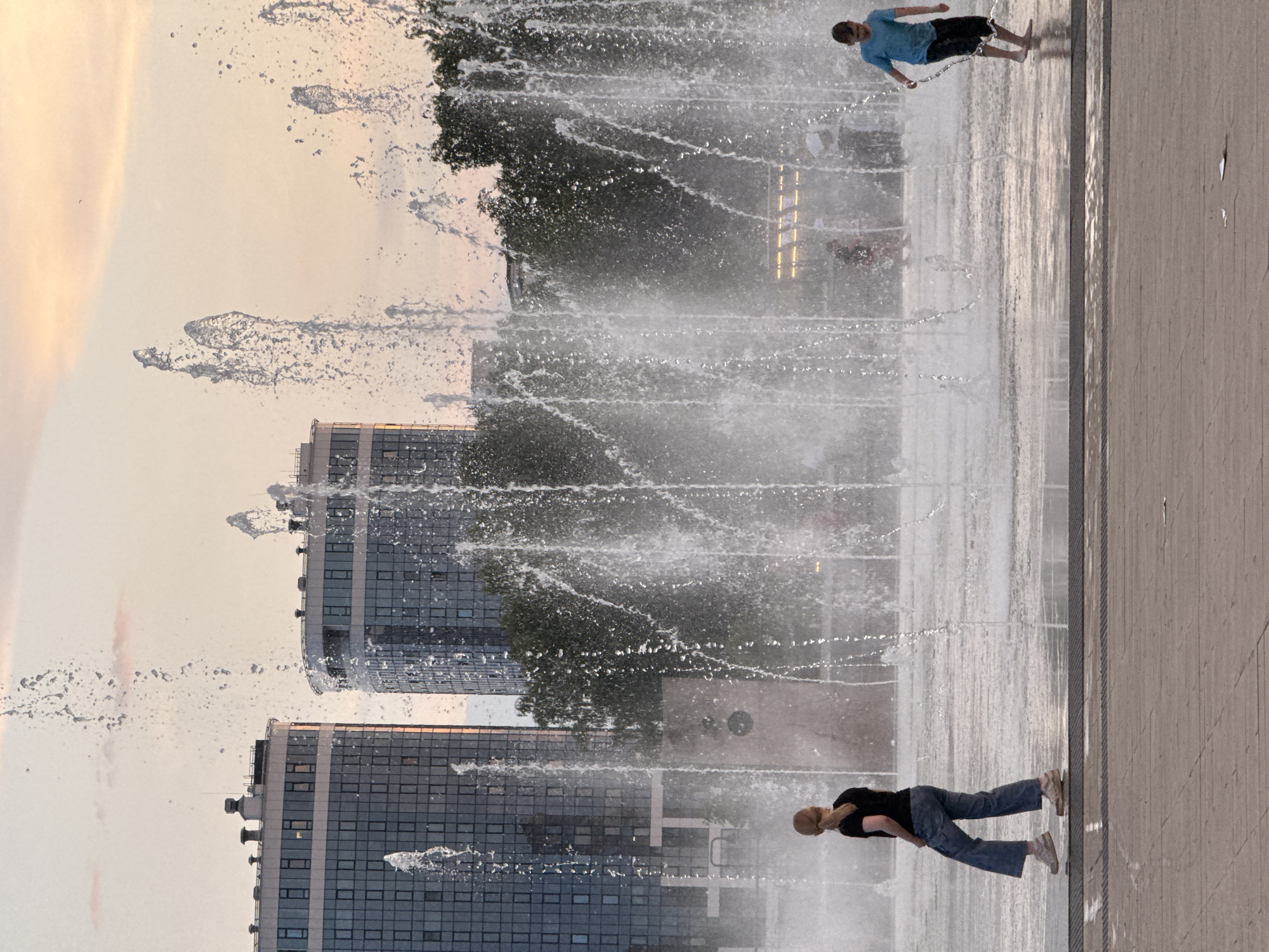 The image depicts a fountain in an urban setting, likely in Izhevsk, Russia, given the context. Two children are visible in the foreground: a girl on the left, wearing a black t-shirt and blue jeans, walks towards the left side of the frame, her hair in a ponytail. On the right, a boy in a blue t-shirt and dark shorts stands, facing towards the left, appearing to be interacting with the water from the fountain. The fountain itself is active, with numerous streams of water shooting upwards and creating a misty effect. In the background, two modern, multi-story buildings with glass facades are prominent, suggesting a commercial or residential area. Trees are interspersed between the buildings and the fountain. The sky is a soft, pale orange and pink, indicative of either sunrise or sunset. The ground in the foreground is paved with large, rectangular concrete slabs.