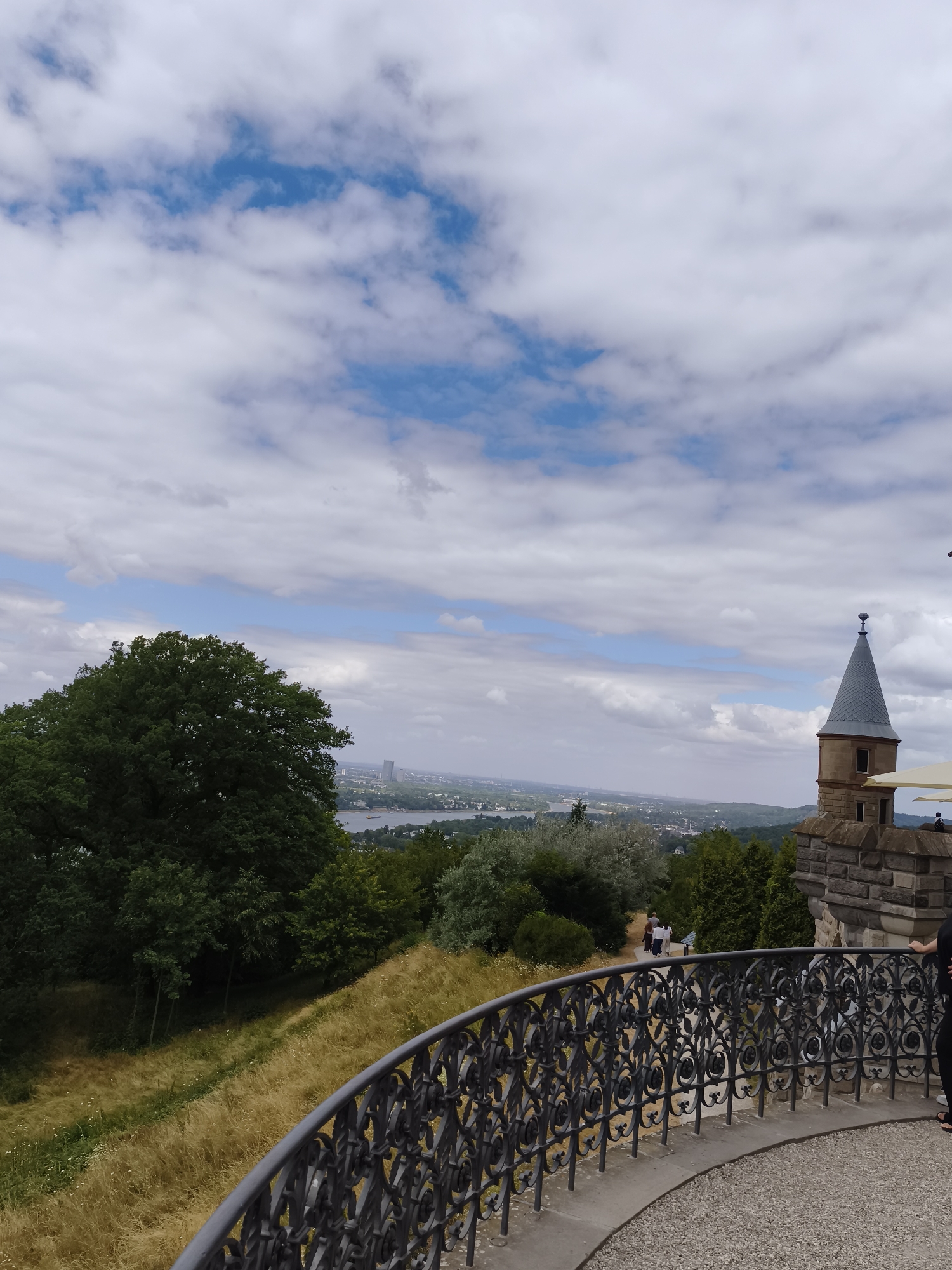 The image captures a panoramic view from the Drachenfels in Königswinter, Germany, on a partly cloudy day. In the foreground, a decorative black metal railing curves along a paved walkway, suggesting a viewpoint or terrace. Beyond the railing, a grassy slope dotted with wild flowers descends towards a lush green forest. Further in the distance, the Rhine River winds through a cityscape, with buildings and boats visible. To the right of the frame, a stone tower with a conical roof, part of the Drachenfels ruins, stands prominently. A few people are visible, one standing near the tower and others walking on a path amidst the greenery. The sky is a mix of blue patches and white clouds, indicative of a dynamic weather system.