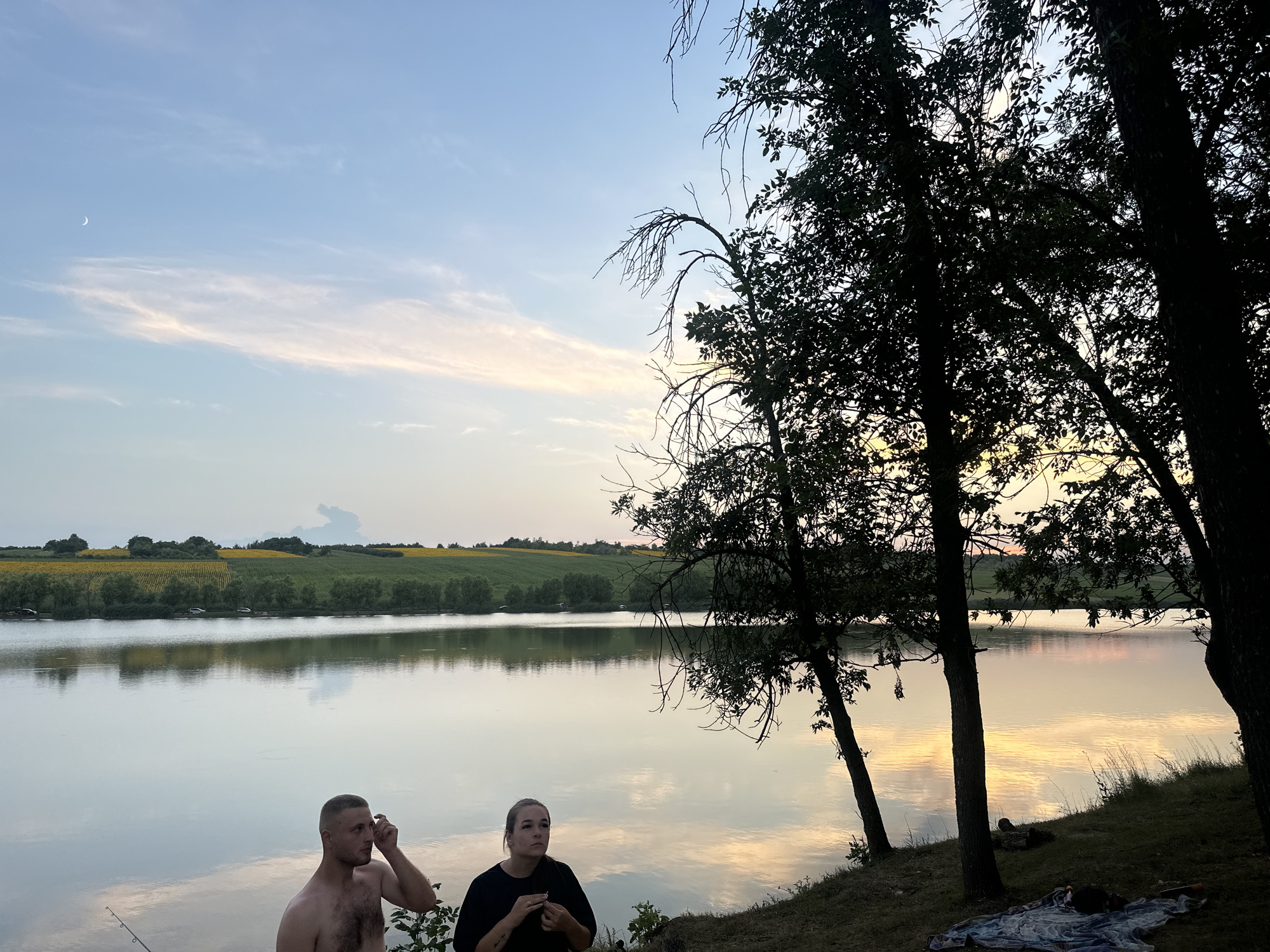 The image depicts a serene outdoor scene at dusk, likely on the outskirts of Găleștii Noi, Moldova. In the foreground, a man with a shaved head, shirtless and with visible chest hair, stands facing right, touching his forehead with his right hand. To his right, a woman wearing a black shirt sits facing left, looking upwards. They are positioned near the edge of a tranquil body of water, with a fishing rod visible to the man's left.
The water reflects the soft hues of the twilight sky, which features scattered clouds tinged with pink and yellow, and a crescent moon visible in the upper left quadrant. Silhouetted against the sky are the dark forms of trees on the right side of the frame, with dense foliage and bare branches.
Beyond the water, a gently rolling landscape is visible, characterized by fields of yellow sunflowers and green crops, bordered by a line of trees. The overall atmosphere is peaceful, capturing a moment of quiet reflection by the water as day transitions into night.