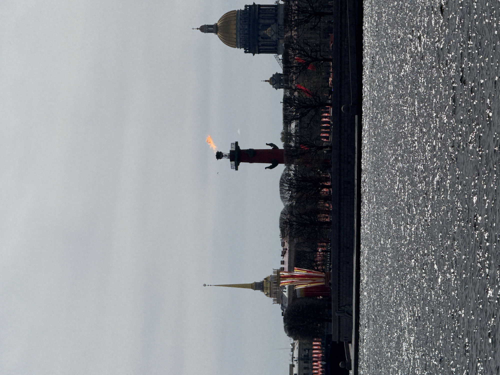 The image captures a view of Saint Petersburg, Russia. Visible are the Rostral Columns with burning flames, Peter and Paul Fortress with its golden spire, and St. Isaac's Cathedral with its golden dome, all situated along the Neva River. The river's surface reflects sunlight. The sky is overcast. Buildings line the river embankment.