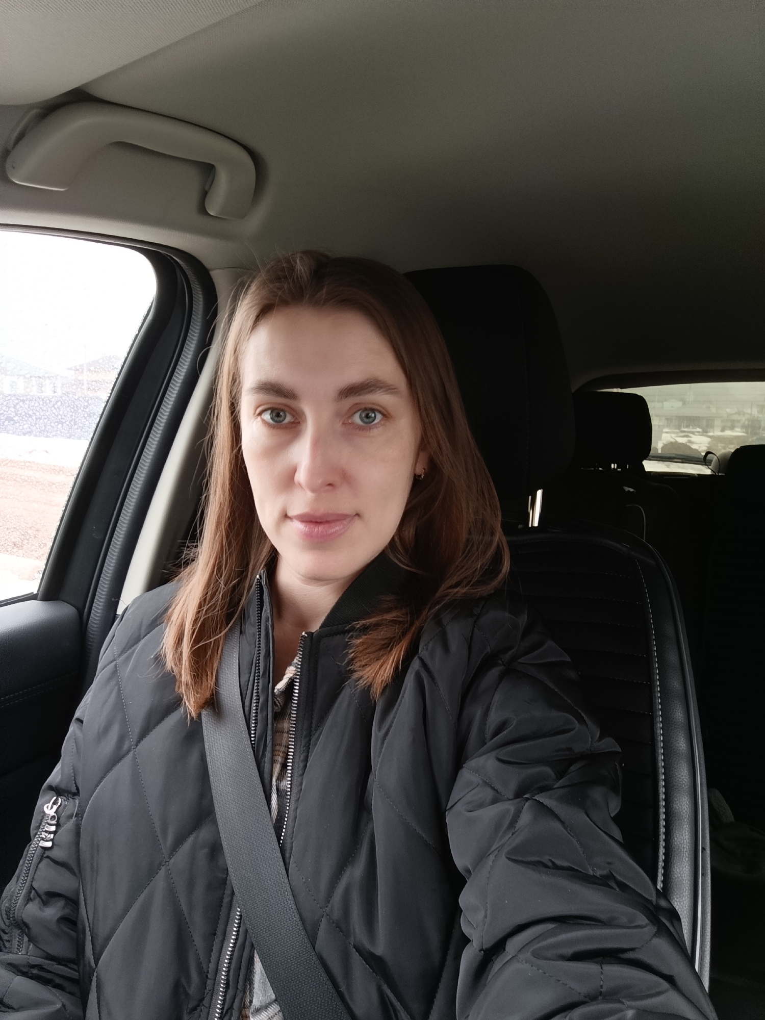 A woman with long, light brown hair and green eyes is seated in the driver's seat of a car, wearing a black quilted jacket and a seatbelt. The background visible through the car windows suggests an urban environment, possibly on the outskirts of a city, with what appears to be low-rise buildings and bare trees. The location is identified as Chaikovsky, Russia.