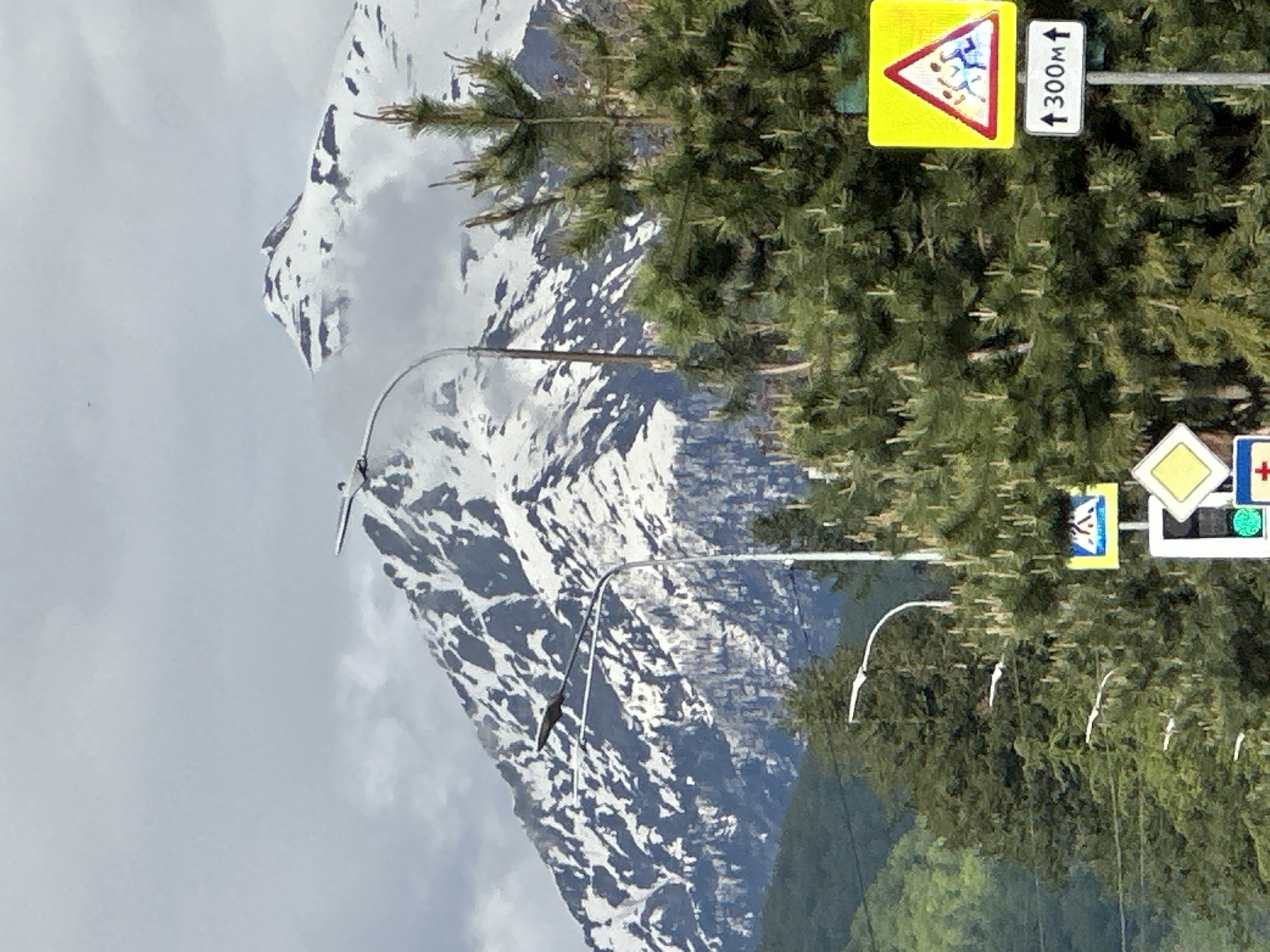 The image captures a view in Krasnaya Polyana, Russia, featuring a snow-covered mountain in the background. In the foreground, there are several road signs, including one indicating a children's area within 300 meters and a pedestrian crossing sign. Streetlights are visible against the backdrop of trees and mountains.