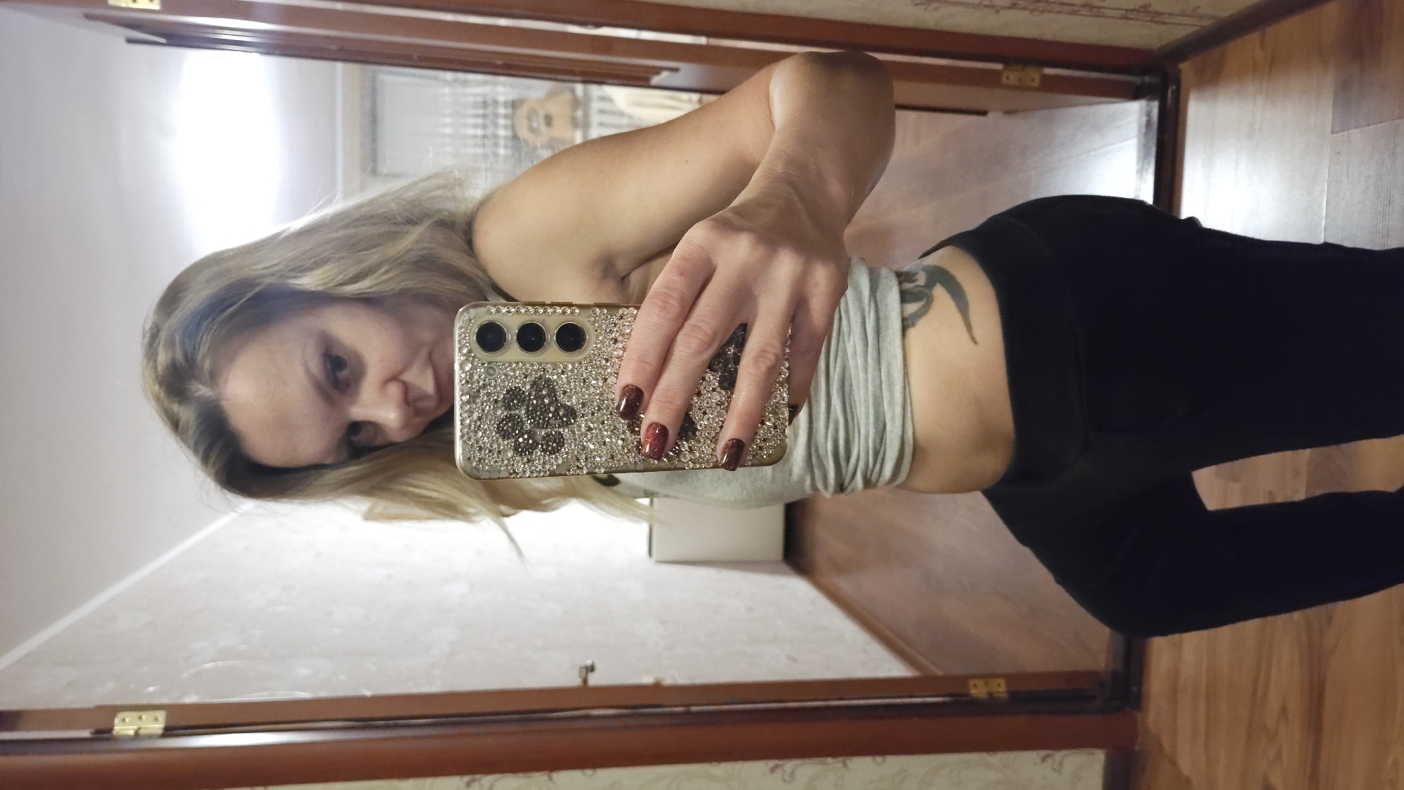 A woman with blonde hair and dark nails is taking a selfie in a mirror. She is wearing a grey top and black leggings. A tattoo is visible on her hip. The environment is an indoor setting with a wooden floor and door frame. The city of Sevastopol, Ukraine is not discernible from the image.