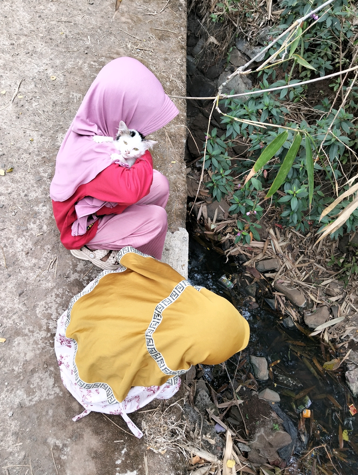 In Cileungsi, Indonesia, a person wearing a pink hijab and a red jacket holds a white and black cat. The person is seated on a concrete surface next to a narrow, shallow stream. Another person, whose lower body is visible, is wearing a yellow garment over a floral-patterned cloth on the ground. The stream is bordered by rocks and overgrown with green vegetation. Debris, including plastic wrappers and dry leaves, is visible in and around the stream. The overall scene depicts an outdoor, informal setting with evidence of litter.