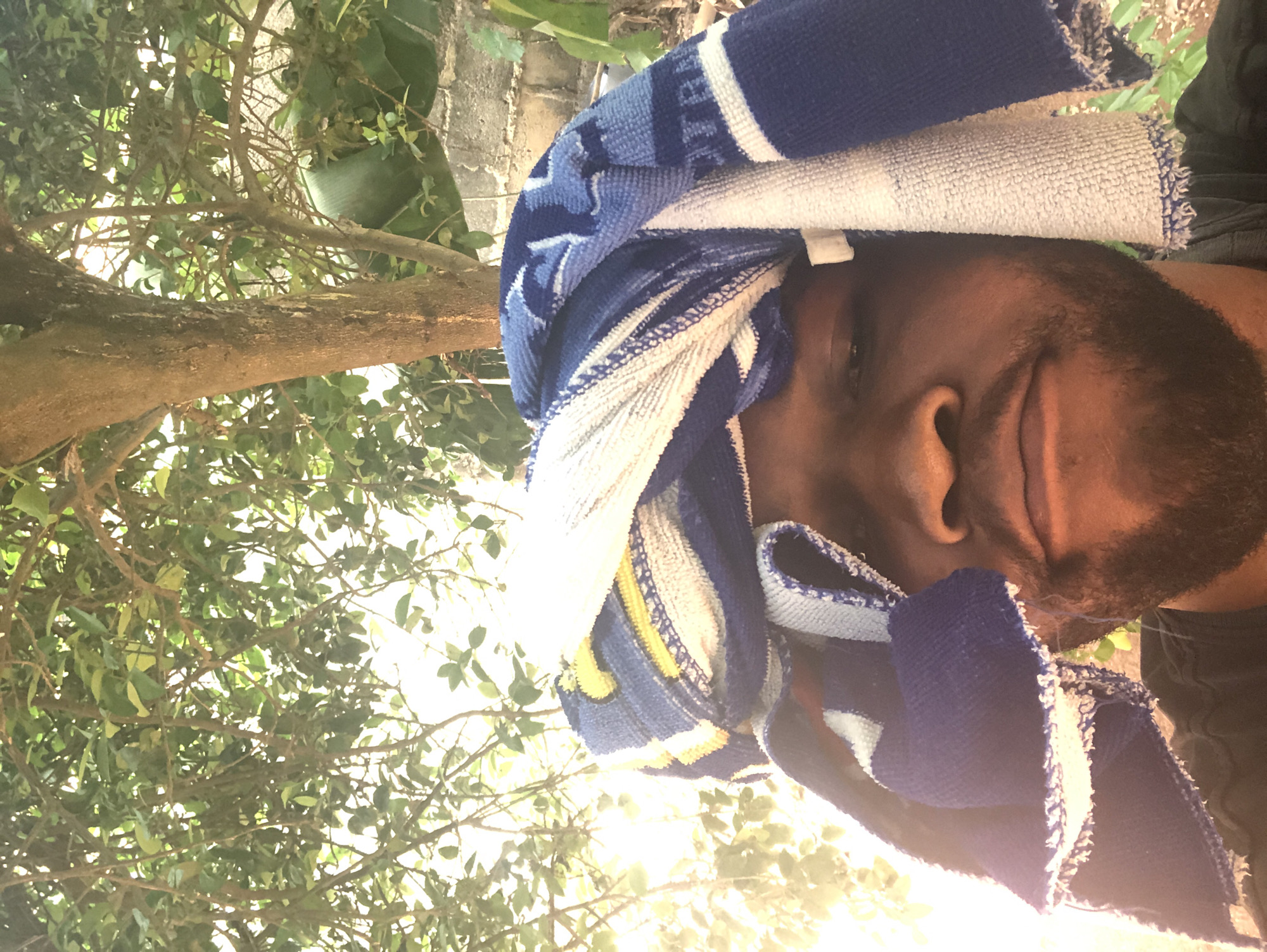 A man is outdoors with trees and greenery in the background. He has a blue and white towel wrapped around his head. The location is in a city of Unknown, Unknown.