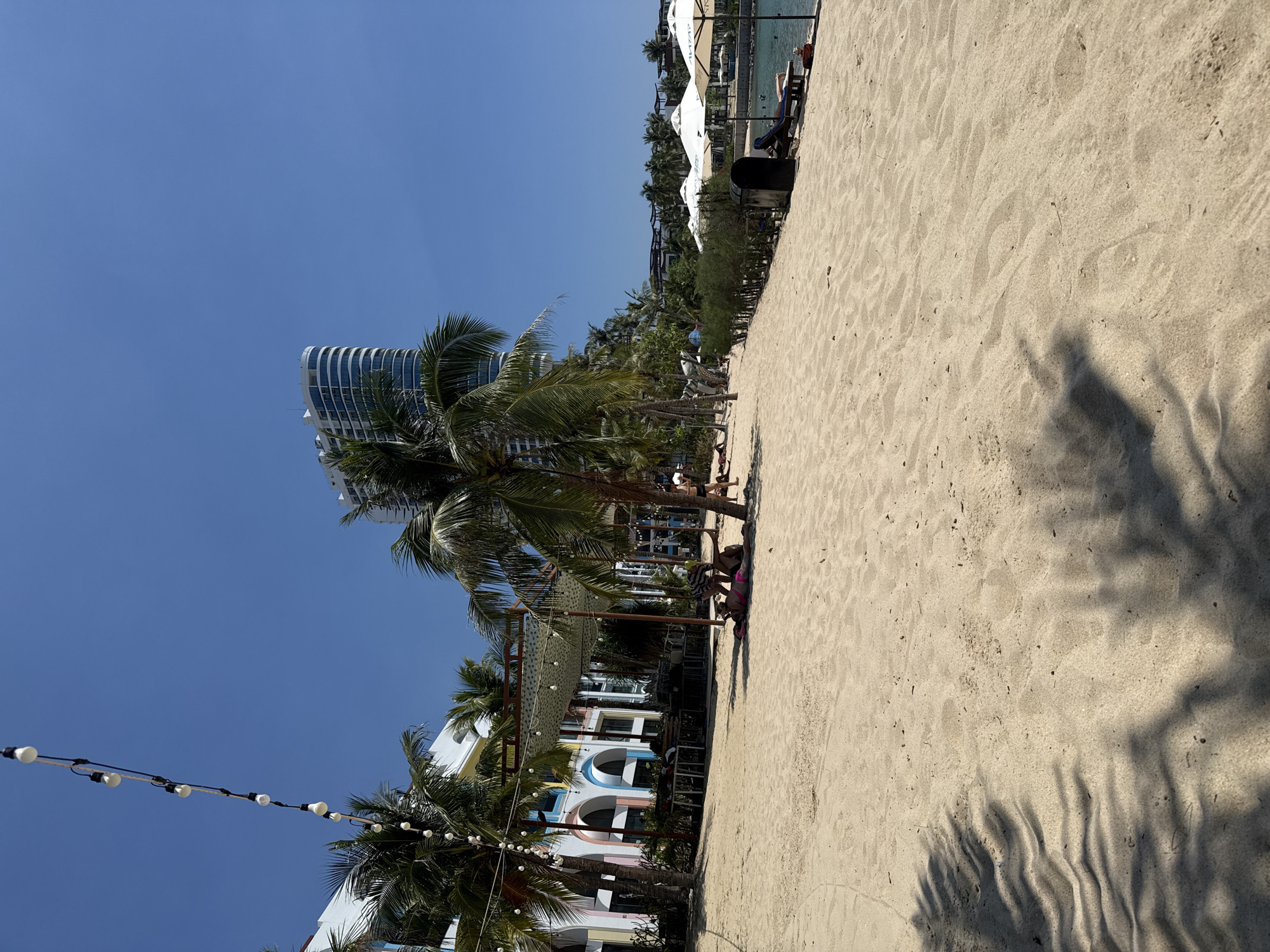 This image depicts a sunny beach scene in Nha Trang, Vietnam. The foreground consists of light-colored sand with visible footprints and rippled textures, marked by dark, elongated shadows from overhead palm fronds.
In the midground, several tall palm trees are present. A string of white globe lights extends diagonally across the upper left quadrant of the sky and continues down, partially strung between palm trees. To the left, a white building with colorful arched windows (light blue, pink, yellow, mint green) is visible, appearing to be a resort structure. In front of this building, under a light-colored awning supported by dark poles, tables and chairs are arranged in an outdoor dining or lounge area, also adorned with string lights.
Immediately to the right of the covered area, a person in a pink bikini lies prone on the sand, partially shaded by a palm tree. Another individual, wearing a striped top, bends over the person in the bikini. Further right and in the background, several people are visible on the sand, some standing, some seated. A row of low green bushes runs parallel to the beach, separating the sand from what appears to be a hard surface or promenade. Beyond the bushes, a line of white beach umbrellas shades lounge chairs situated directly next to calm, blue-green water. One umbrella distinctly displays "AVATA" in dark lettering. A dark trash bin is positioned near the umbrellas.
The background features multiple high-rise buildings, including a prominent white, curved skyscraper with numerous windows, located towards the center-right. Additional greenery and more buildings line the distant shore. The sky is clear and intensely blue.