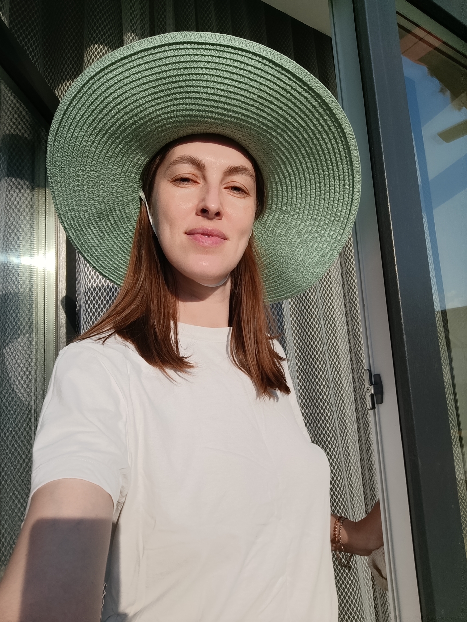A woman wearing a light green wide-brimmed hat and a white t-shirt stands in front of a door with a screen. The door frame is gray, and reflections can be seen in the glass. The screen is of a grid pattern. Part of her arm with bracelets is visible in the lower right corner. The location is likely Chaikovsky, Russia.
