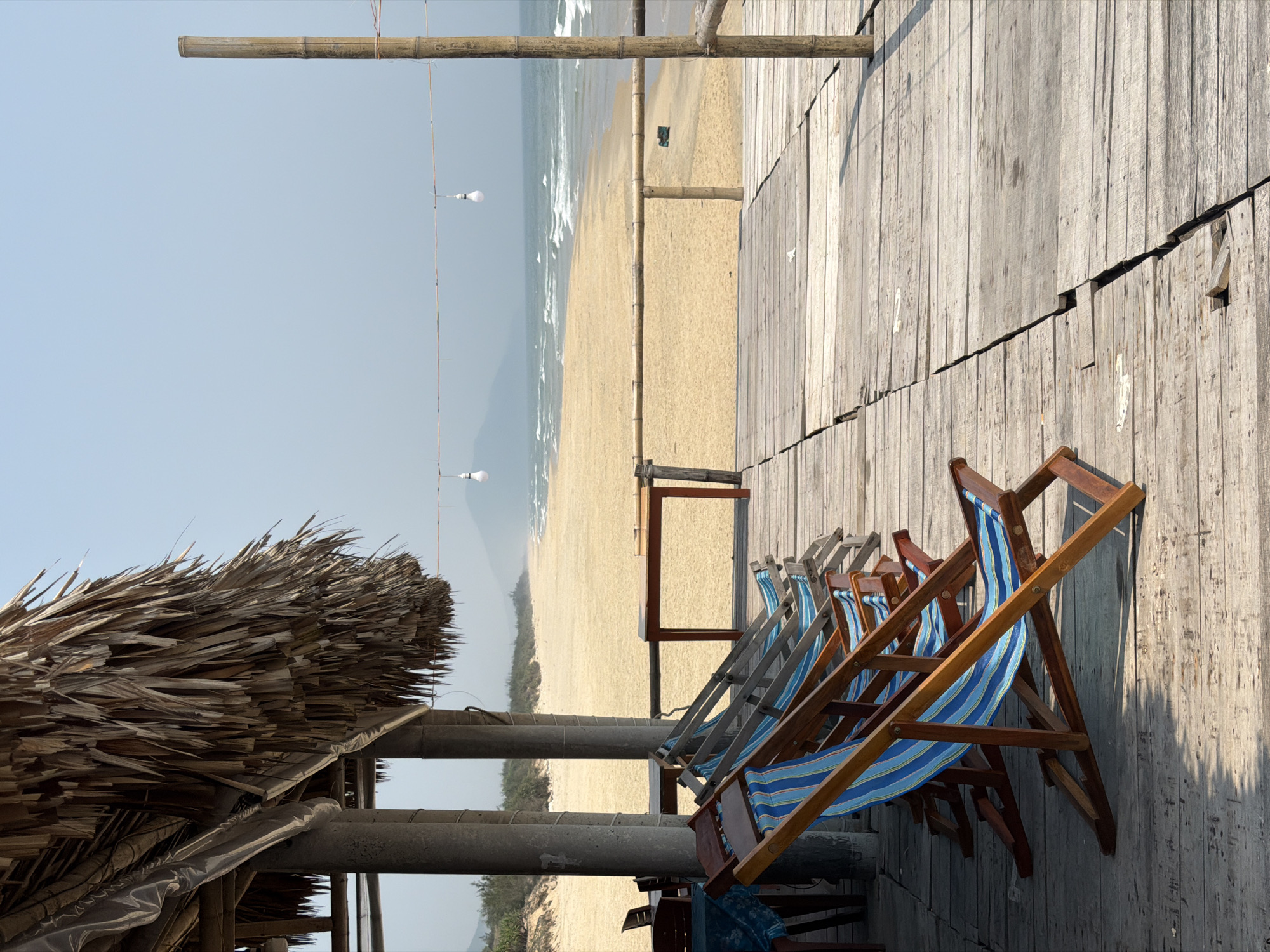 The image captures a beach scene, likely taken in a coastal area near Huế, Vietnam. A wooden deck with several blue and white striped beach chairs is visible in the foreground. The deck leads to a sandy beach with ocean waves and a clear sky in the background. Overhead, bamboo poles support a series of hanging light bulbs. A thatched roof structure stands on the left side of the frame. The photograph is taken from a low angle, emphasizing the texture and details of the wooden deck and beach chairs.