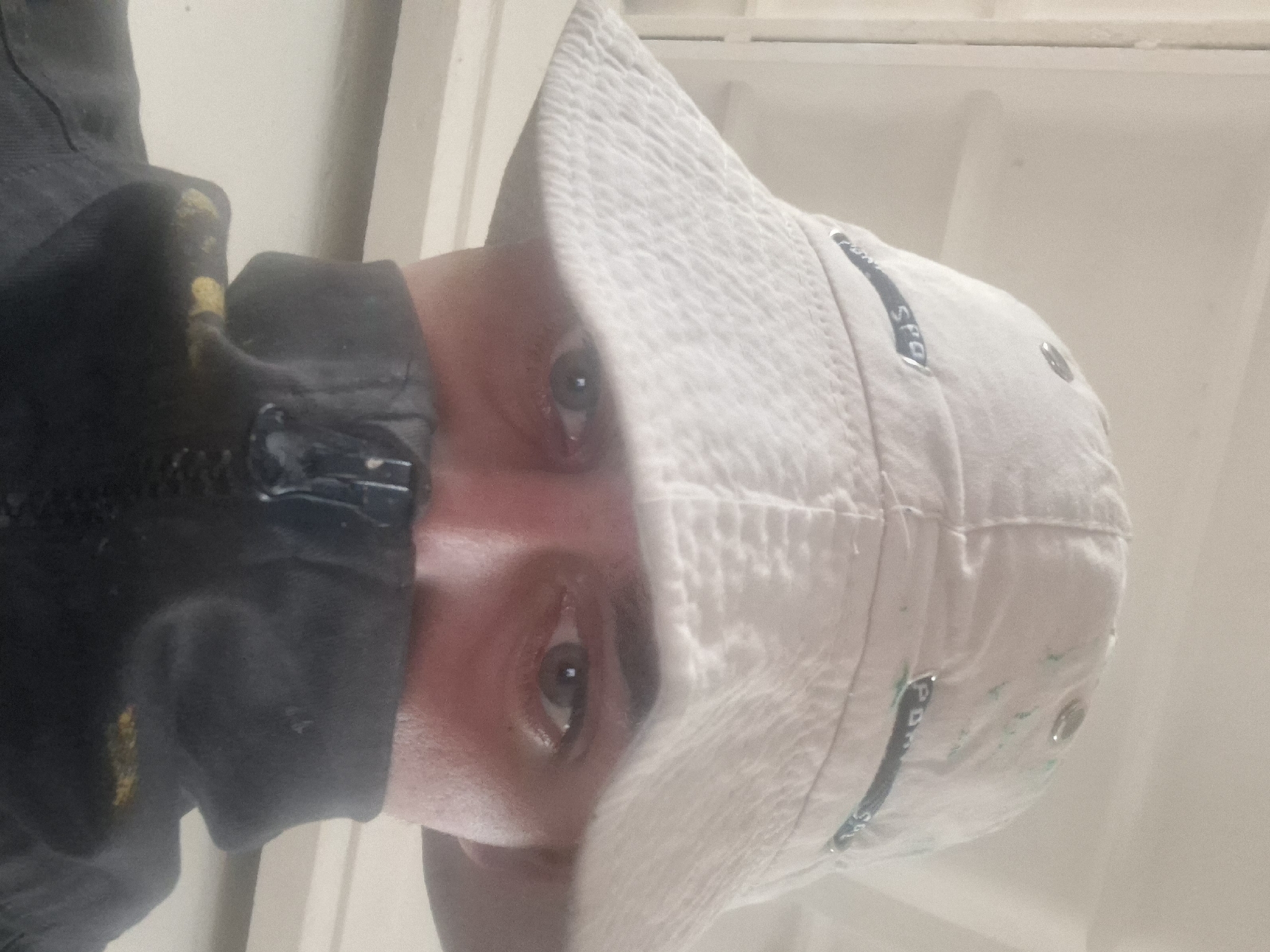 A medium close-up shows a person wearing a white bucket hat with dark blue accents and metal details on the right side. The hat is slightly stained with green. The person's face, only partially visible, shows brown eyes and pinkish skin. They are wearing a black jacket with a partially visible zipper. The background is white and indeterminate. The location is Unknown, Unknown.