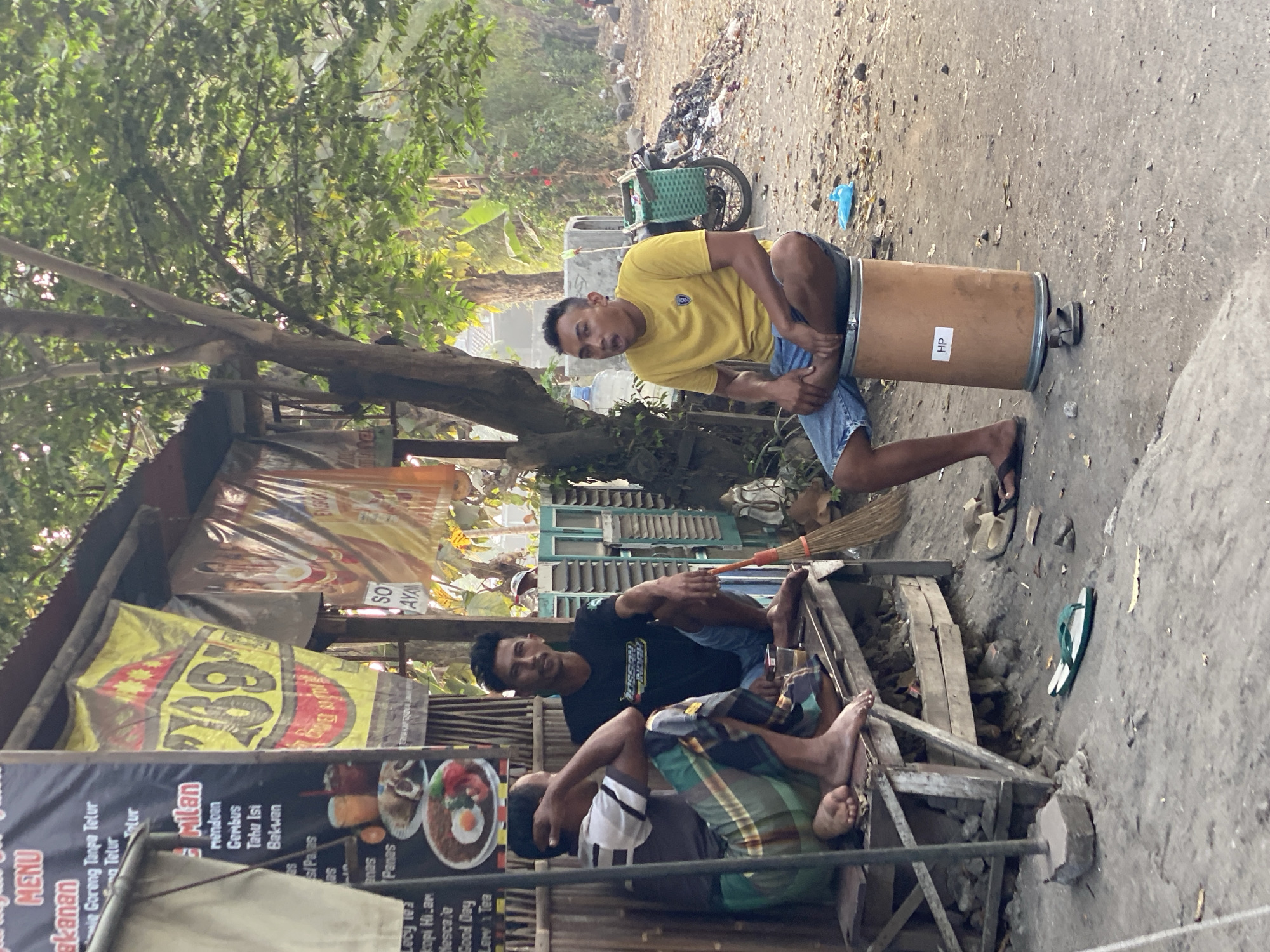 In Kasihan, Indonesia, three men are gathered outside a roadside food stall. Two men are seated on a wooden bench, while the third man sits on a large, cylindrical drum. The stall features a tin roof and bamboo walls, adorned with various colorful banners advertising food items. One banner displays the number "289" prominently. The ground around the stall is dirt and gravel, with some debris and a bicycle with a green basket visible in the background. The man on the drum is wearing a yellow t-shirt and blue shorts, and he appears to be speaking. The other two men are dressed casually, with one wearing a black t-shirt and the other a checkered sarong. A broom leans against the bench, and a pair of flip-flops are scattered on the ground. The overall scene depicts a casual outdoor gathering at a local eatery.