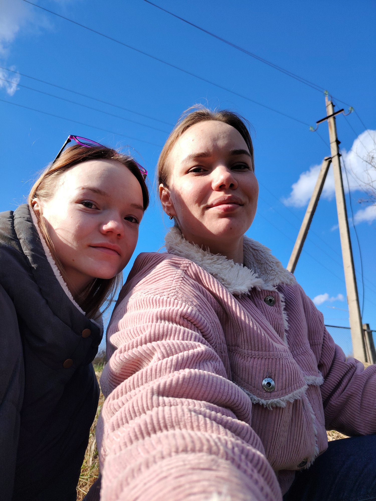 Two young women are posed in an outdoor selfie on a sunny day. The woman on the left wears a dark jacket with a light-colored lining and sunglasses perched on her head. The woman on the right wears a pink corduroy jacket with a fuzzy white collar. Behind them, a clear blue sky is visible, along with utility poles and power lines. The location appears to be in a rural or suburban area within the city of Chaikovsky, Russia, based on the type of utility poles and the general environment.