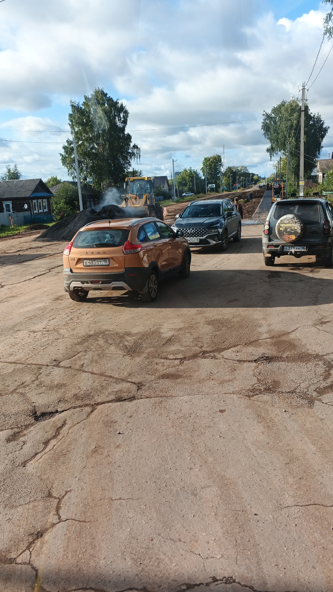 In the rural settlement of Kiyasovo, Russia, a street construction scene unfolds under a cloudy sky. The foreground is dominated by a cracked asphalt surface, indicative of ongoing roadwork. In the middle ground, an orange Lada car is parked on the dirt road. Behind it, a dark SUV, possibly a Hyundai, is also stopped. Further down the road, a dark grey SUV, identified as a Lada Niva, is visible. The backdrop features a yellow excavator actively engaged in moving dirt, releasing a plume of dust. Residential buildings, typical of a rural Russian village, are scattered amongst trees on either side of the road. Utility poles and wires stretch across the scene, typical of rural infrastructure. The overall atmosphere suggests a typical day of infrastructure improvement in a small Russian town.