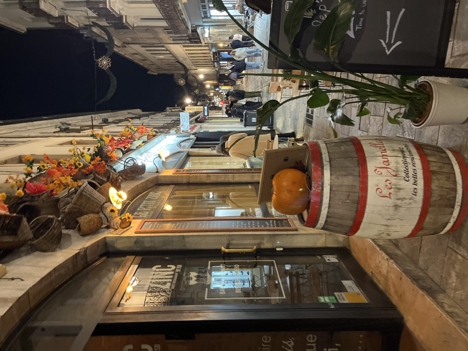 A nighttime street scene shows a European-style street lined with buildings decorated for autumn. A barrel is decorated with the words "Les Jamelly" and holds a pumpkin in a cardboard box. A chalkboard with an arrow pointing down is next to the barrel. "LE PETIT ZINC" is displayed above a window. People stroll in the distance, under soft lighting. Decorative lights and floral arrangements adorn the building facades, and potted plants add to the charm.