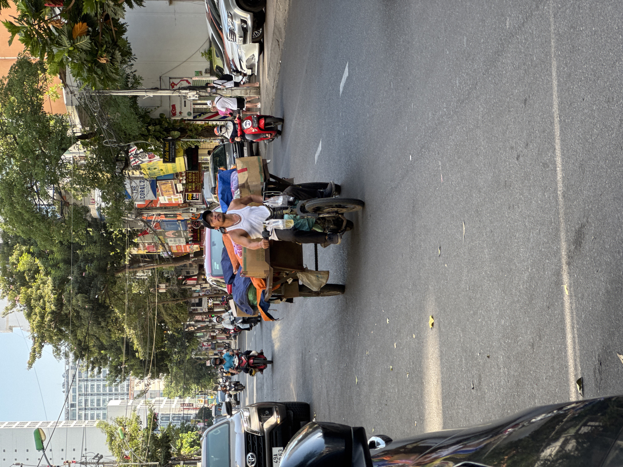 The image depicts a street scene in Nha Trang, Vietnam, with a focus on a man riding a motorcycle that is pulling a cart laden with various goods. The man, wearing a white tank top and dark pants, is positioned in the foreground, looking ahead. The cart is a two-wheeled trailer attached to the rear of his motorcycle. It is piled high with what appear to be textiles, including blue, orange, and brown fabrics, as well as cardboard boxes.
The street is paved and shows faint white lane markings. Several other vehicles are visible on the road, including cars and motorcycles. To the left, a dark-colored SUV is prominent, and further down the street, a red motorcycle with a rider is also visible. In the background, buildings line the street, some featuring signage with Vietnamese text. Tall trees with dense green foliage arch over the roadway, providing shade. The sky is clear and bright. The overall impression is that of a busy urban street with active commerce and transportation.