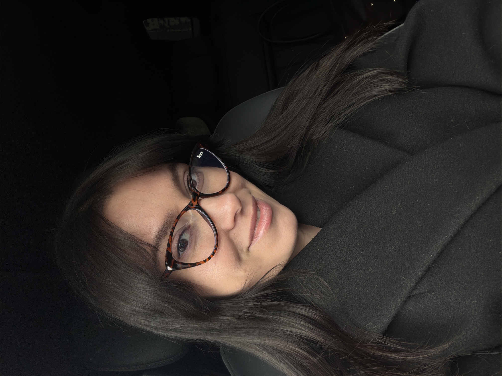 A woman with long, dark hair and glasses is shown in a close-up, slightly angled selfie. She is wearing a dark-colored coat or jacket. The background is dark and indistinct, but suggests she is inside a car. There is no information to confirm the location is Izhevsk, Russia.