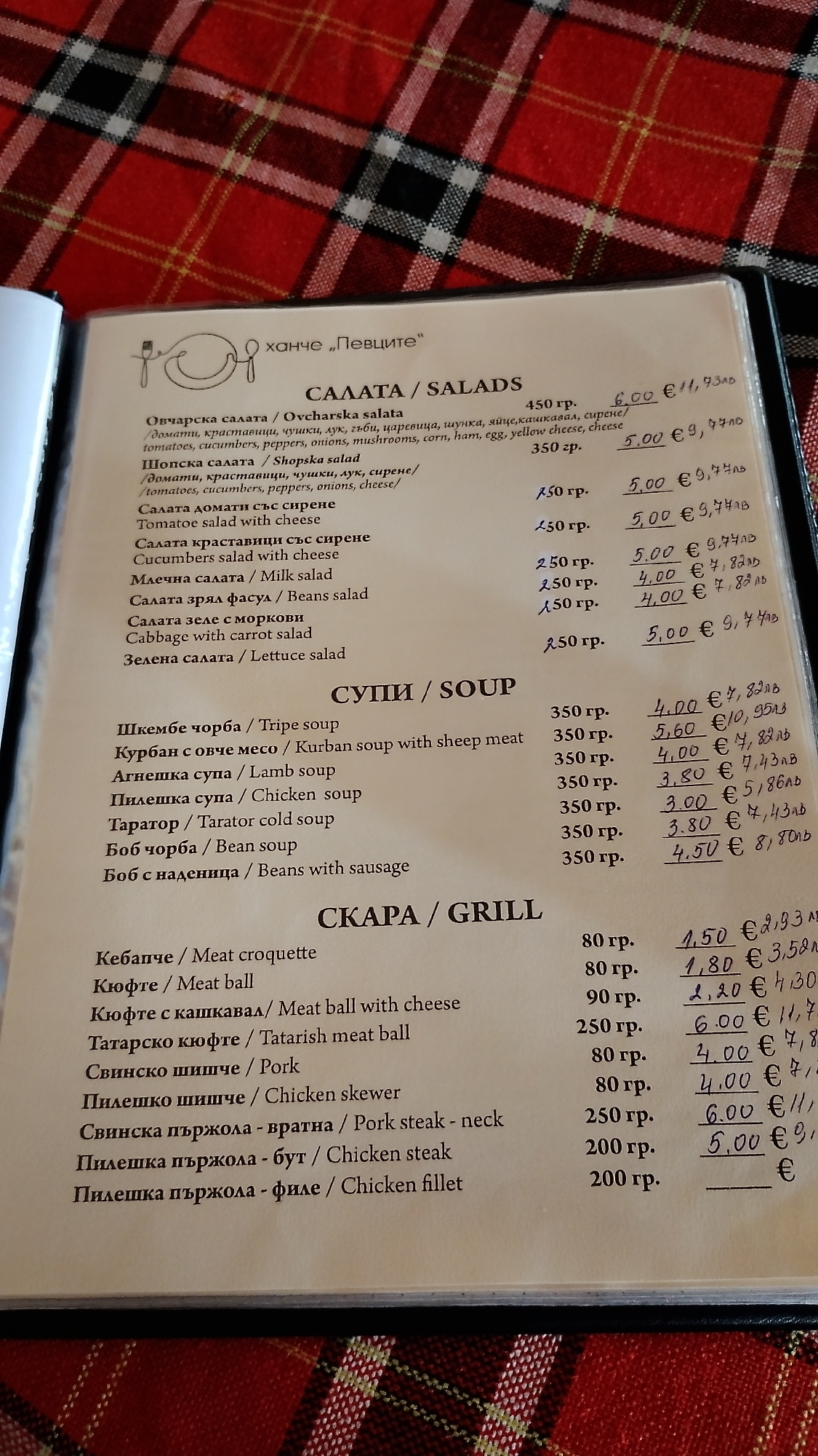 The image shows a menu from a restaurant named "Ханче „Певците“" (Hanched "Pevtsite"), likely located in Pevtsite, Bulgaria. The menu is presented in Bulgarian and English, divided into sections for Salads, Soups, and Grill. The items listed include various salads like Ovcharska and Shopska, soups such as Tripe and Lamb soup, and grill items like meat croquettes and chicken skewers. Prices are listed in Bulgarian Lev (лв) and Euros (€). The menu is open on a table with a red and black plaid tablecloth visible in the background, suggesting an indoor setting.