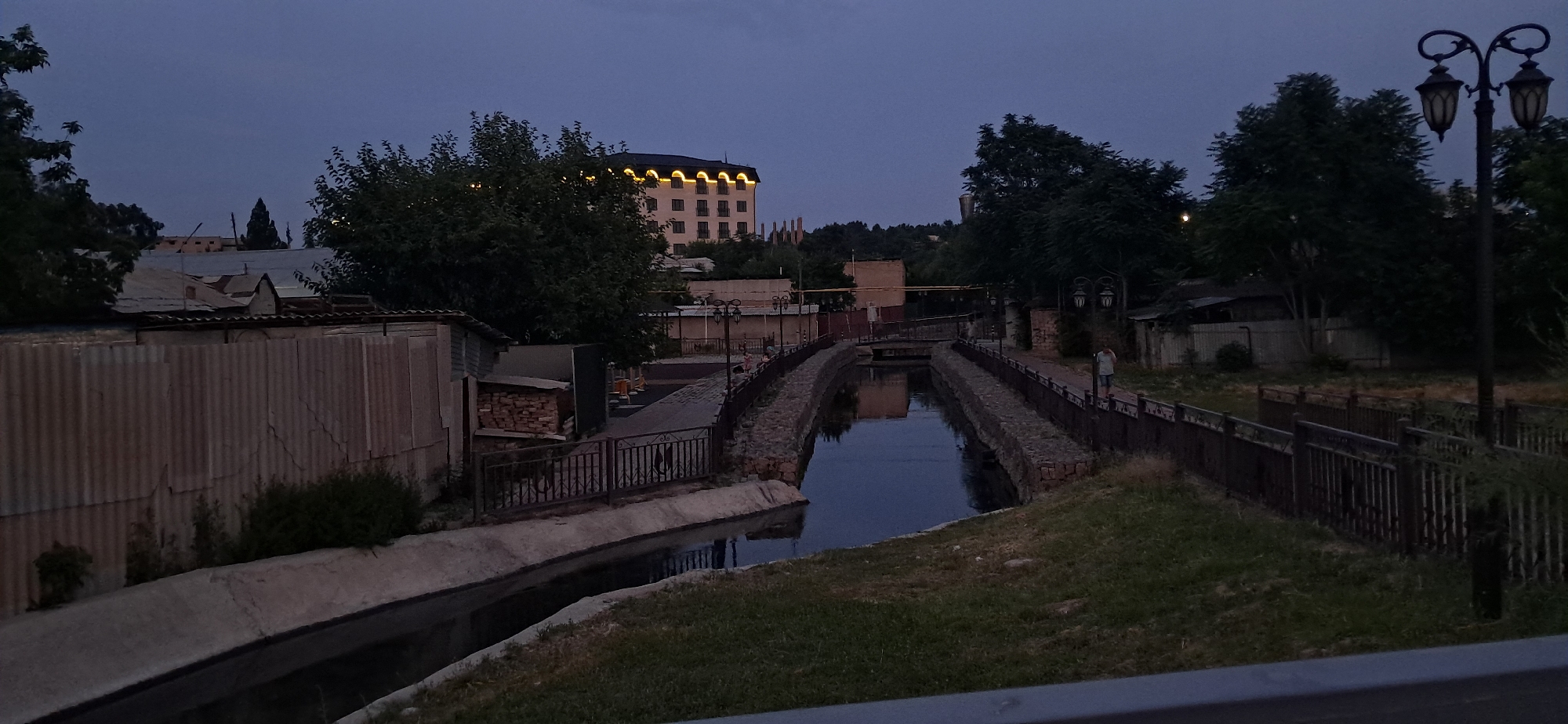 In the city of Unknown, Unknown, at dusk, a view is captured of a canal lined with stone walls and a metal fence. A single person is walking on the path next to the canal. On the left side of the canal, there is a corrugated metal fence and some low-lying structures. Across the canal, a building with lights along its roofline is visible, surrounded by trees. The sky is a deep blue, indicating the transition from day to night. Several decorative lampposts are spaced along the path.